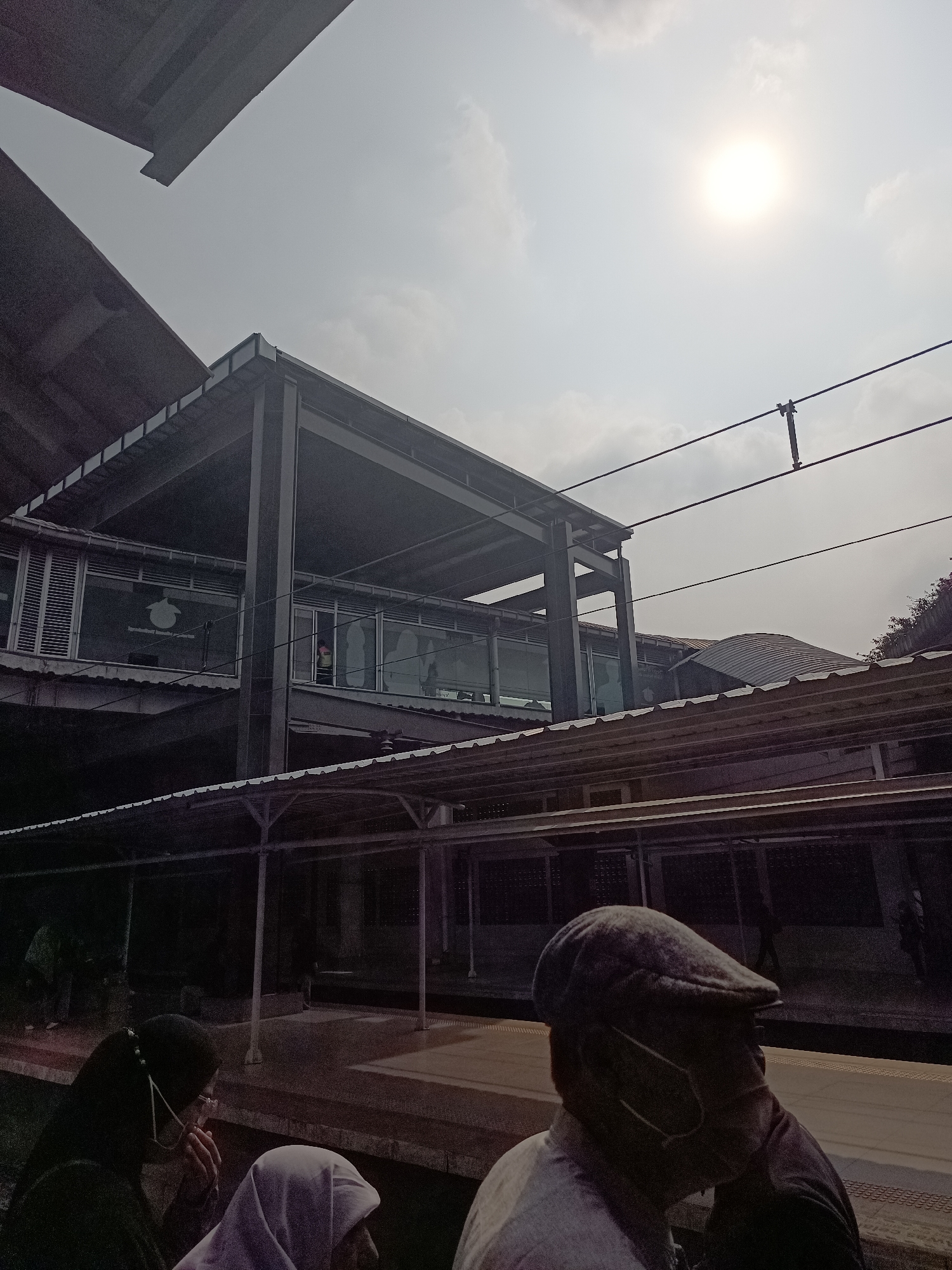 The image depicts a view of a modern train station in Central Jakarta, Indonesia, under a bright, overcast sky. The foreground features the back of a person wearing a flat cap and a mask, their face turned to the right. To their left, two individuals in headscarves are visible, one in dark clothing and the other in light purple, also wearing masks. They appear to be waiting on a platform.
The station itself has a multi-level structure with a dark, metal frame supporting covered walkways and platforms. Large glass windows are visible on the upper levels, reflecting the sky and interior lights. Overhead, electrical wires stretch across the frame, indicating the presence of an electrified railway system. The overall impression is of a functional and contemporary public transportation hub.