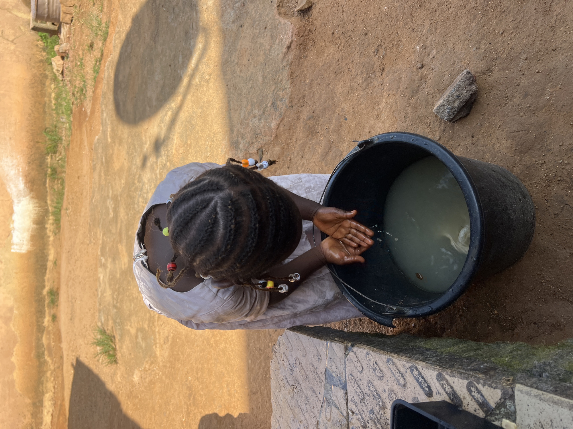 A young Black female is washing her hands in a large black bucket of water. She is wearing a light-colored dress and has her hair braided with beads. The ground is sandy dirt, and there are some small green plants. In the background, there is a wall of a building made of a rough, tan material. A shadow of a person is cast on the ground. The scene appears to be outdoors in a somewhat rustic setting. This location is consistent with areas in Jos, Nigeria, where water access can be challenging and simple washing stations are common.