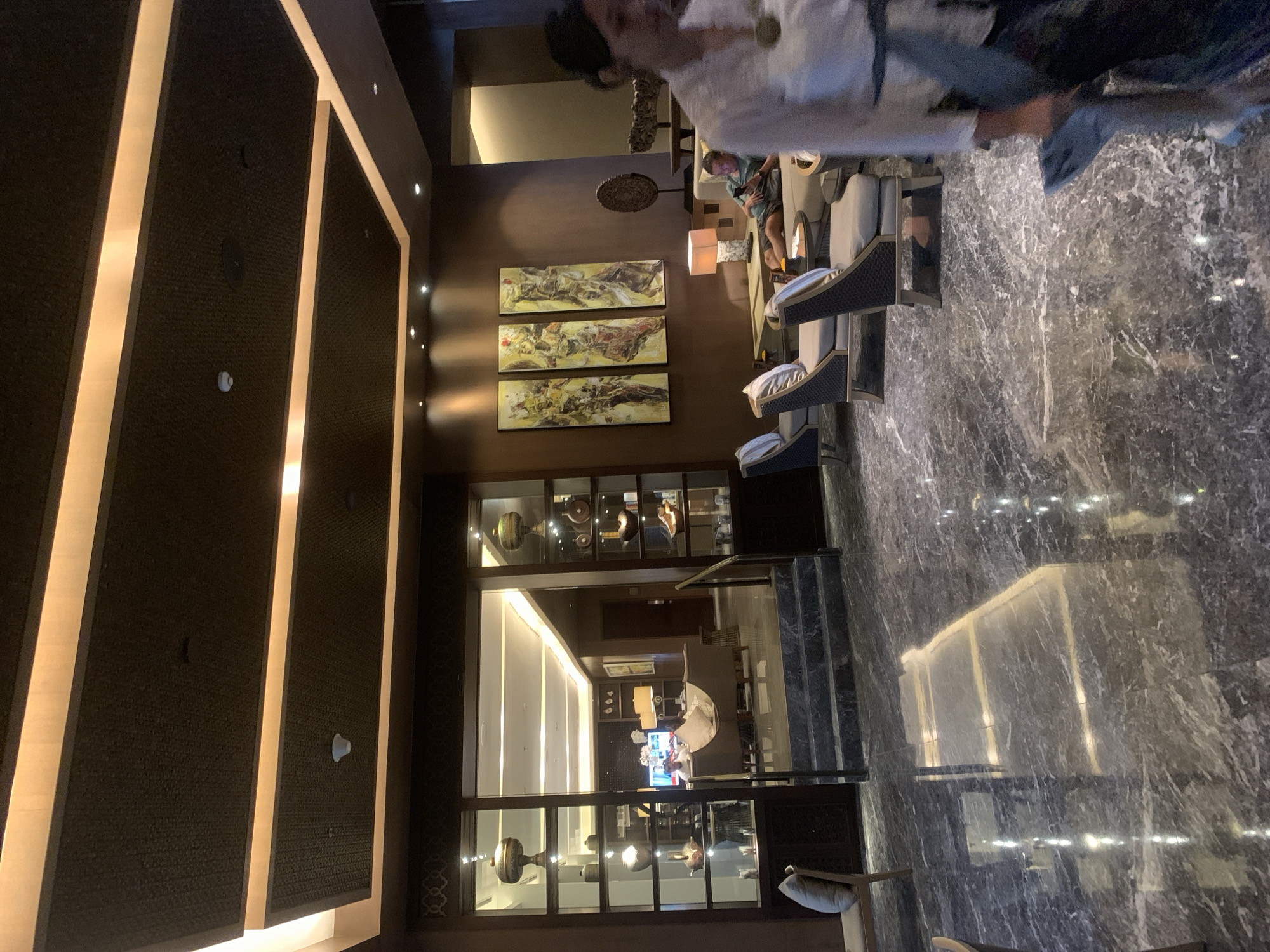 The image depicts the interior of a hotel lobby in Kuta, Indonesia. The scene is characterized by luxurious furnishings and decor. The floor is made of polished dark marble with white veining, reflecting the ambient lighting. In the foreground, a person in a white shirt and dark pants is walking away from the camera. Further back, a man in a green t-shirt and shorts is seated on a sofa, looking at his phone. The lobby features comfortable seating areas with upholstered chairs and sofas. On the wall, three framed artworks depicting figures in yellow and brown tones are displayed. The ceiling has a dark textured finish with recessed lighting strips. A large glass-fronted display cabinet filled with decorative vases and objects is visible on the left side of the image, along with a staircase leading to a higher level. The overall ambiance suggests a sophisticated and well-appointed hotel lobby.
