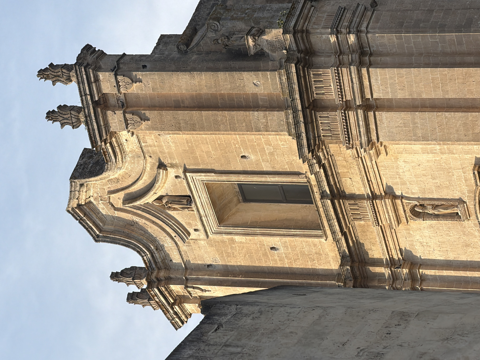 The image shows the facade of a historic building in Matera, Italy, likely a church or palazzo. The architecture features intricate stone carvings, a large rectangular window, and decorative elements such as finials and statues. The building is constructed of light-colored stone blocks and appears to be bathed in the warm light of the setting sun, casting long shadows. A portion of a lighter-colored, rough-textured wall is visible to the left of the main facade. The sky above is clear and a pale blue. The scene is static and depicts an architectural detail without any discernible human activity.
