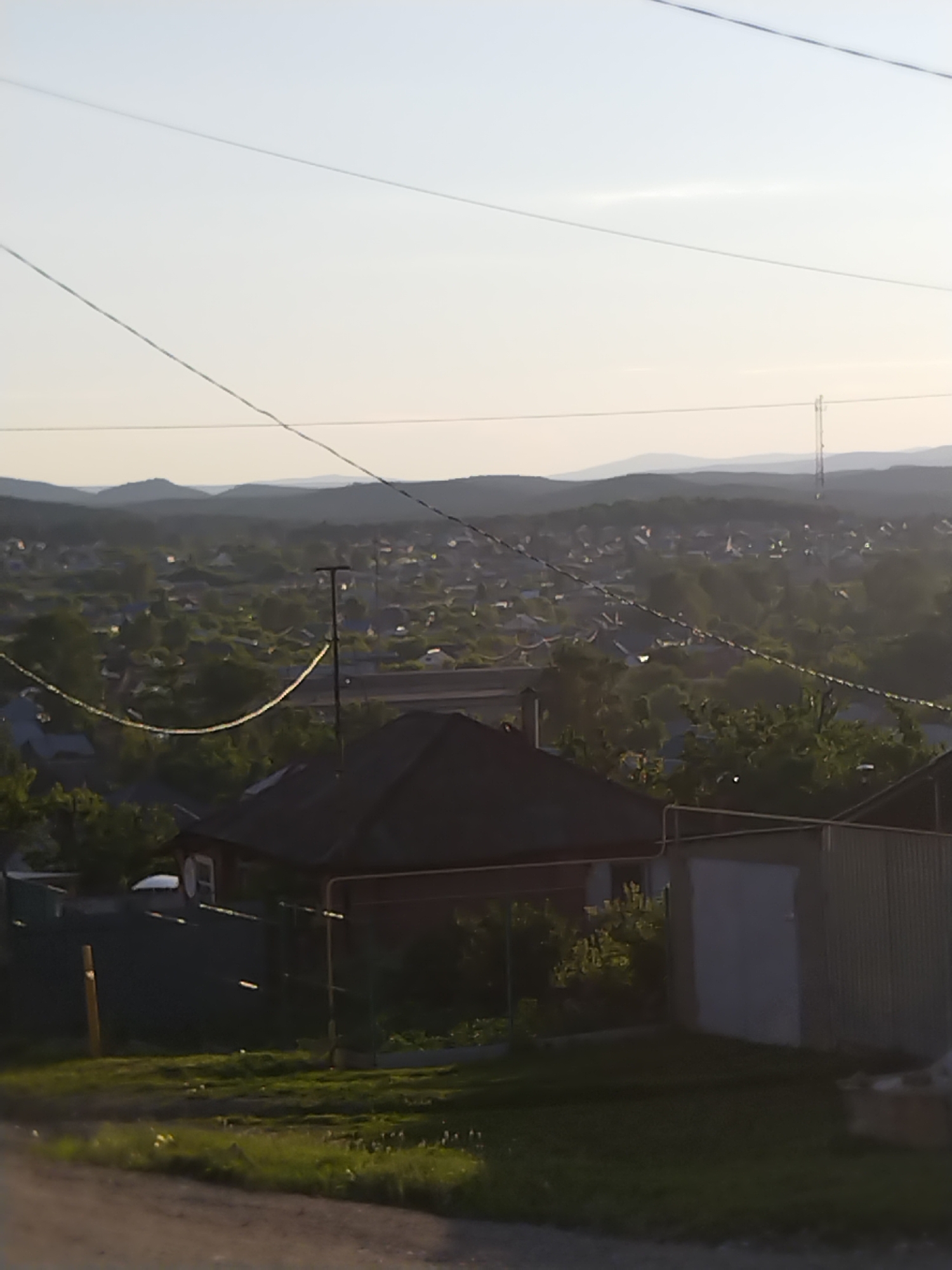 The image depicts a panoramic view of a rural village nestled amidst rolling hills. The scene is bathed in the soft, warm light of either sunrise or sunset, casting long shadows and creating a serene atmosphere. Overhead, power lines stretch across the frame, adding a touch of human infrastructure to the natural landscape.
In the foreground, the roofs of several houses are visible, with one prominent dark-roofed house in the lower center of the image. The village itself is densely populated with houses of varying sizes and styles, interspersed with lush green trees. The distant hills form a layered backdrop, fading into a hazy sky.
The time of day is suggested by the low angle of the sun and the warm, diffused light. The weather appears clear, with no clouds visible in the sky. The location is likely a country or village setting, characterized by the presence of individual homes, trees, and the vast expanse of undeveloped land. There is no visible text in the image. The overall impression is one of quietude and natural beauty, with a hint of human habitation in harmony with the environment.