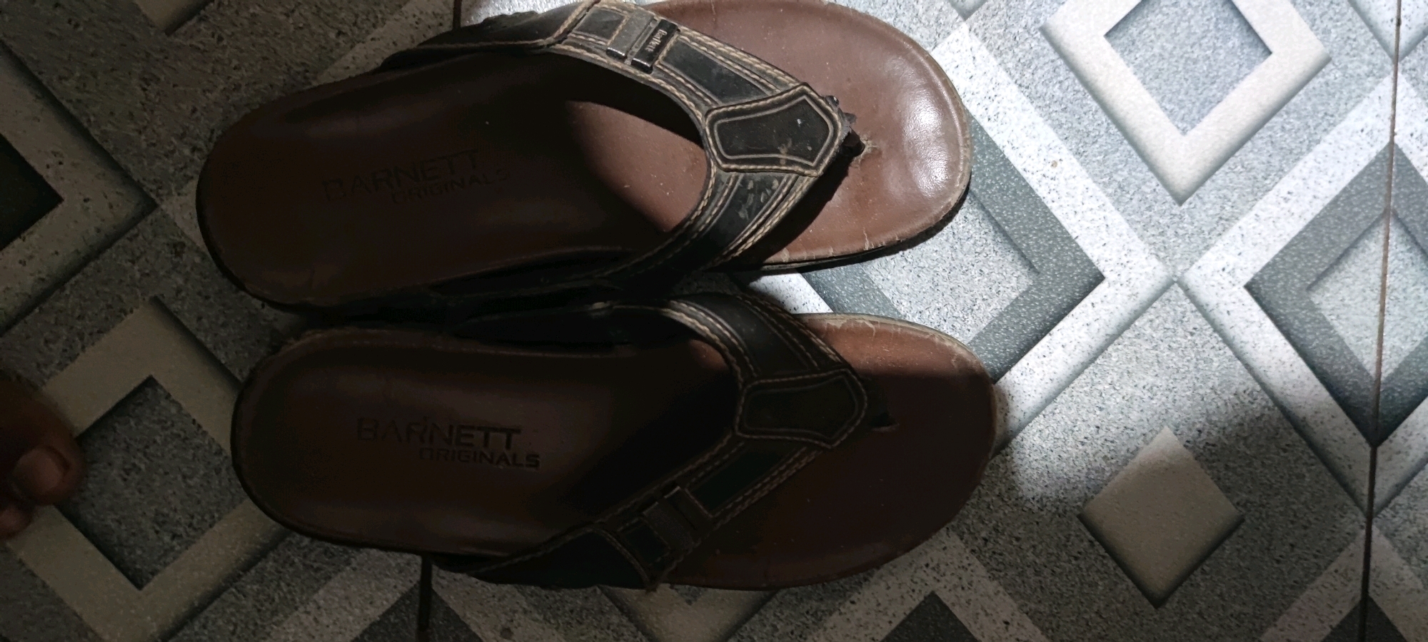 A pair of brown Barnett Originals flip-flops are placed on a patterned tiled floor in Gambarsari, Indonesia. The flip-flops are made of leather with black straps and stitching. The left flip-flop is on top of the right one. The floor tiles are a diamond pattern with shades of gray and white. The lighting in the image is dim, casting shadows on the flip-flops and the floor.