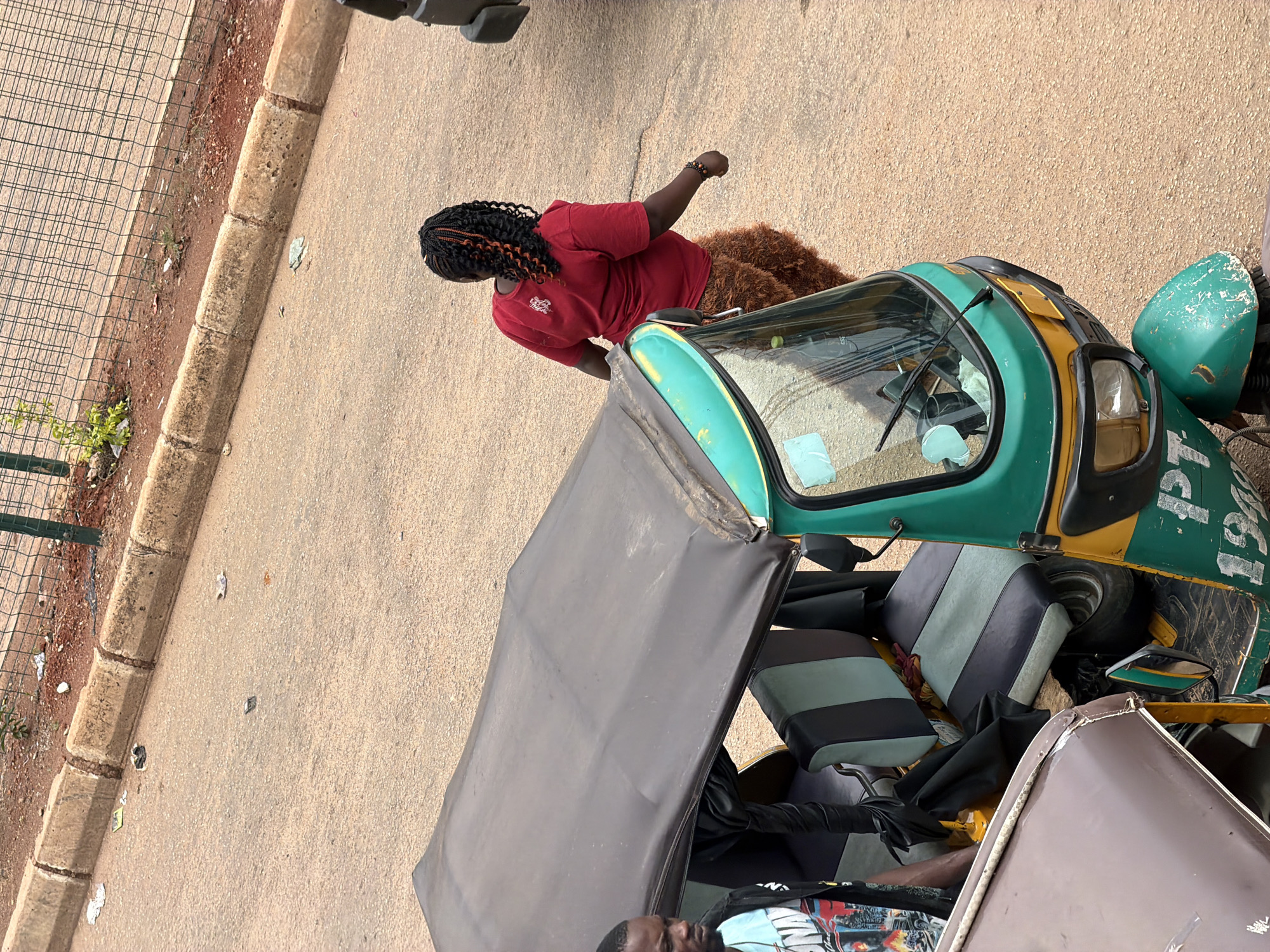 In Bukuru, Nigeria, an overhead shot captures a busy street scene. A woman with braided hair, wearing a red t-shirt and brown shaggy pants, walks away from the camera. She is near a green and yellow auto-rickshaw, its dark canopy extended. A man with a beard, wearing a blue t-shirt with a graphic print, stands next to the auto-rickshaw, looking towards it. The auto-rickshaw has striped seating and appears to be parked on a paved road. The environment is dusty, with a concrete curb and a wire fence in the background, suggesting an urban or semi-urban setting. Litter is visible on the ground, indicating a common urban condition.