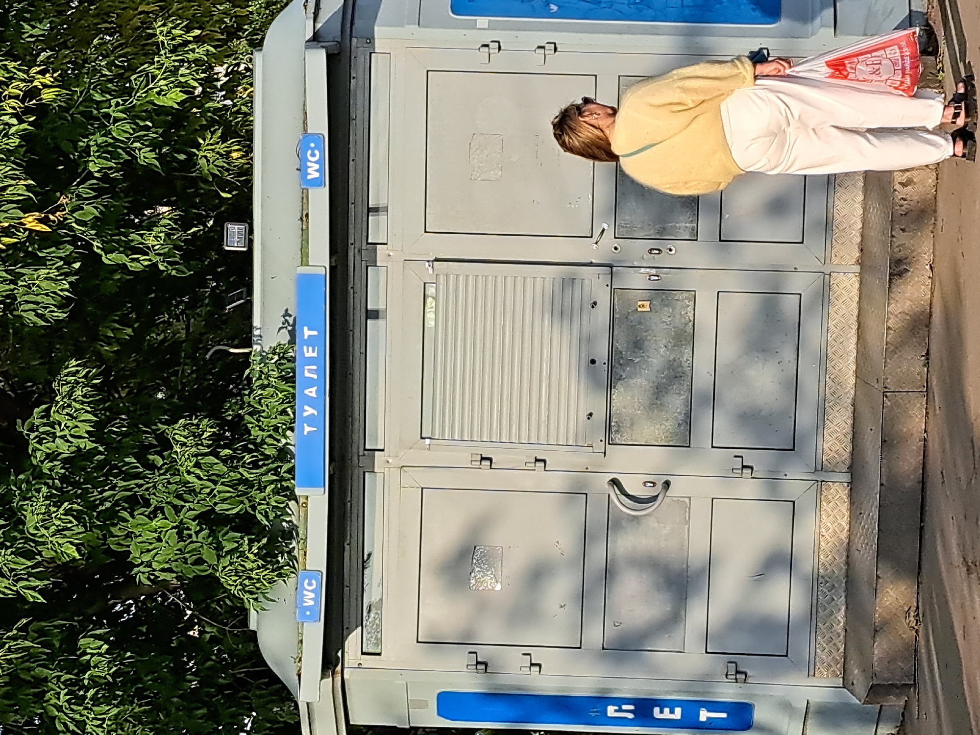 The image shows a woman standing in front of a public toilet facility in Irkutsk, Russia. The toilet is a gray, prefabricated structure with multiple doors and blue signage indicating "WC" and "ТУАЛЕТ" (Toilet). The woman is wearing a yellow sweater and white pants, and she is holding a red and white plastic shopping bag. The background consists of green trees and a blue sky. The sun is casting shadows on the ground, suggesting it is daytime. The overall scene depicts an urban environment with a functional public amenity.