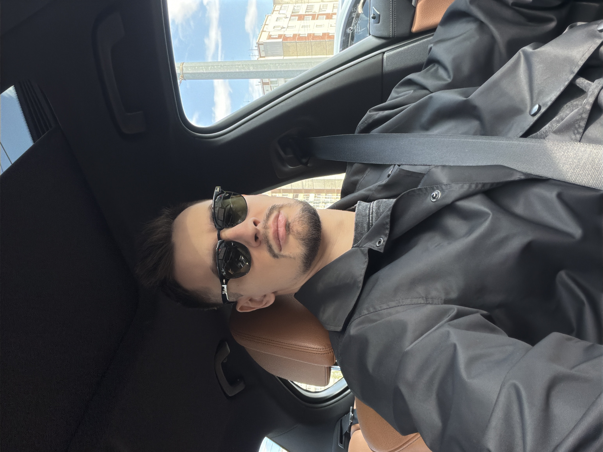 A man wearing sunglasses and a black jacket is seated in a car. The car has a sunroof that shows a building in the background and a blue sky. The interior of the car has brown leather seats. The location is Krasnoyarsk, Russia. The man is wearing a seatbelt.