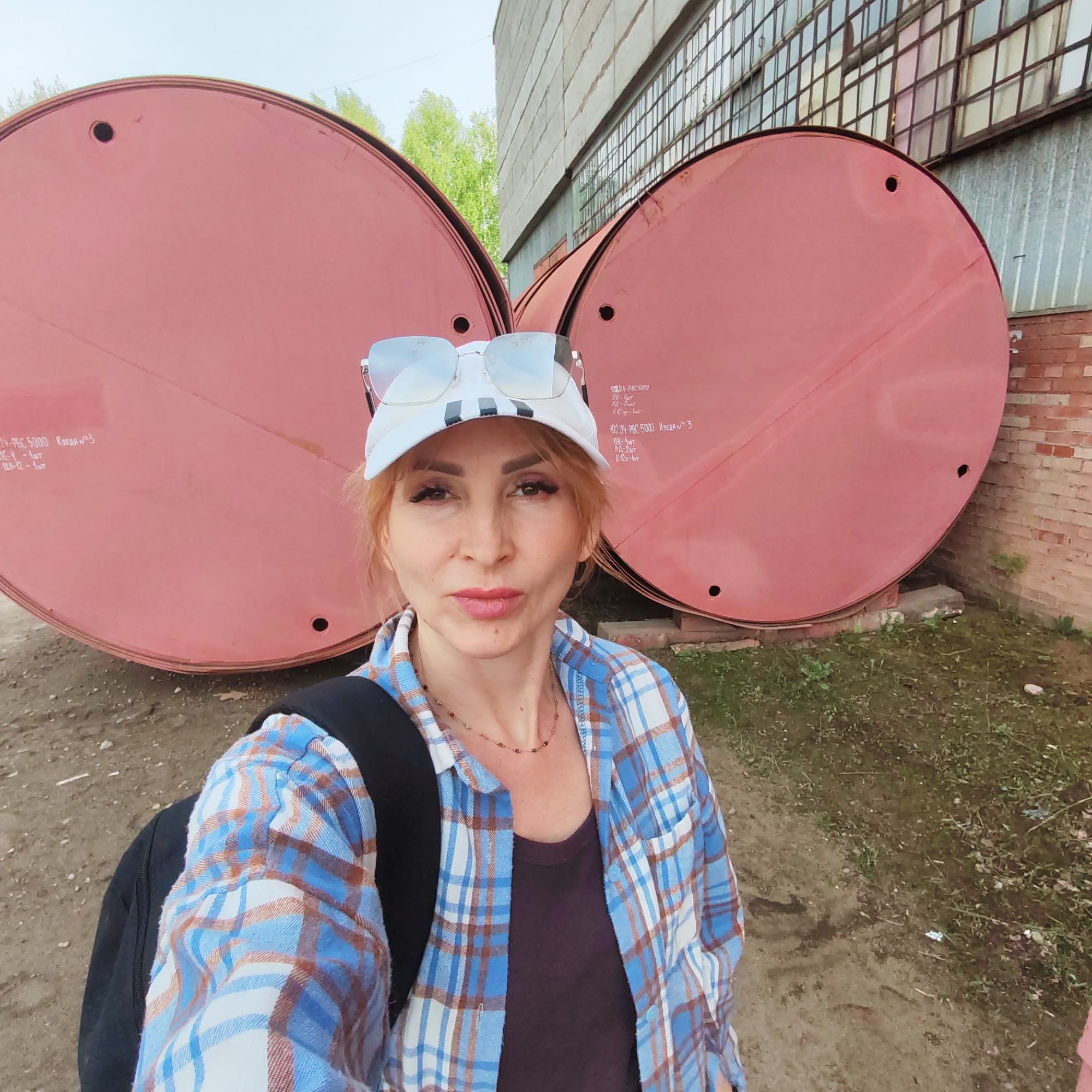 A woman with auburn hair, wearing a blue and brown plaid shirt, a dark purple shirt, a necklace, and a white baseball cap with sunglasses resting on top, poses for a selfie in an industrial area. She has a black backpack slung over her shoulder. Behind her are two large, round, pink tanks, each with small openings near the top. The tanks appear to be placed against a brick wall and a large, windowed industrial building. The ground is dirt and patchy grass. Inscriptions are visible on the surface of the tanks. The scene is located in the city of Chaikovsky, Russia.