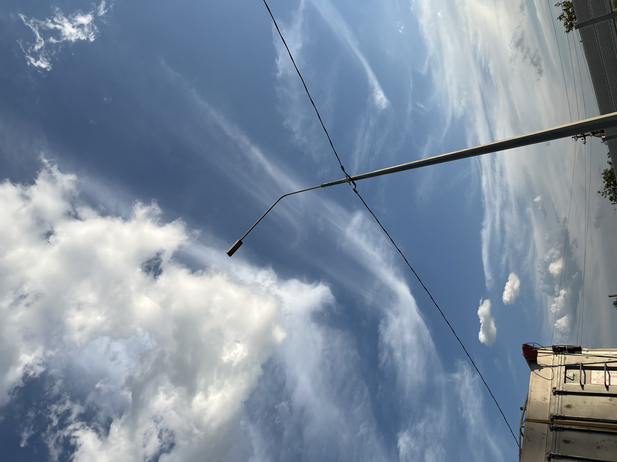 The image depicts a bright, daytime scene with a clear blue sky dominated by various cloud formations. Wispy cirrus clouds stretch across the sky, interspersed with larger, fluffier cumulus clouds. A single street lamp pole stands diagonally in the foreground, extending from the bottom right towards the top left of the frame. A dark wire runs horizontally across the middle of the image, attached to the street lamp pole. In the lower left corner, a portion of a metallic structure, possibly a building or vehicle, is visible. In the lower right, a grey, textured surface resembling a fence or wall is present, with some greenery visible behind it. The overall impression is an outdoor, possibly rural or suburban, environment.