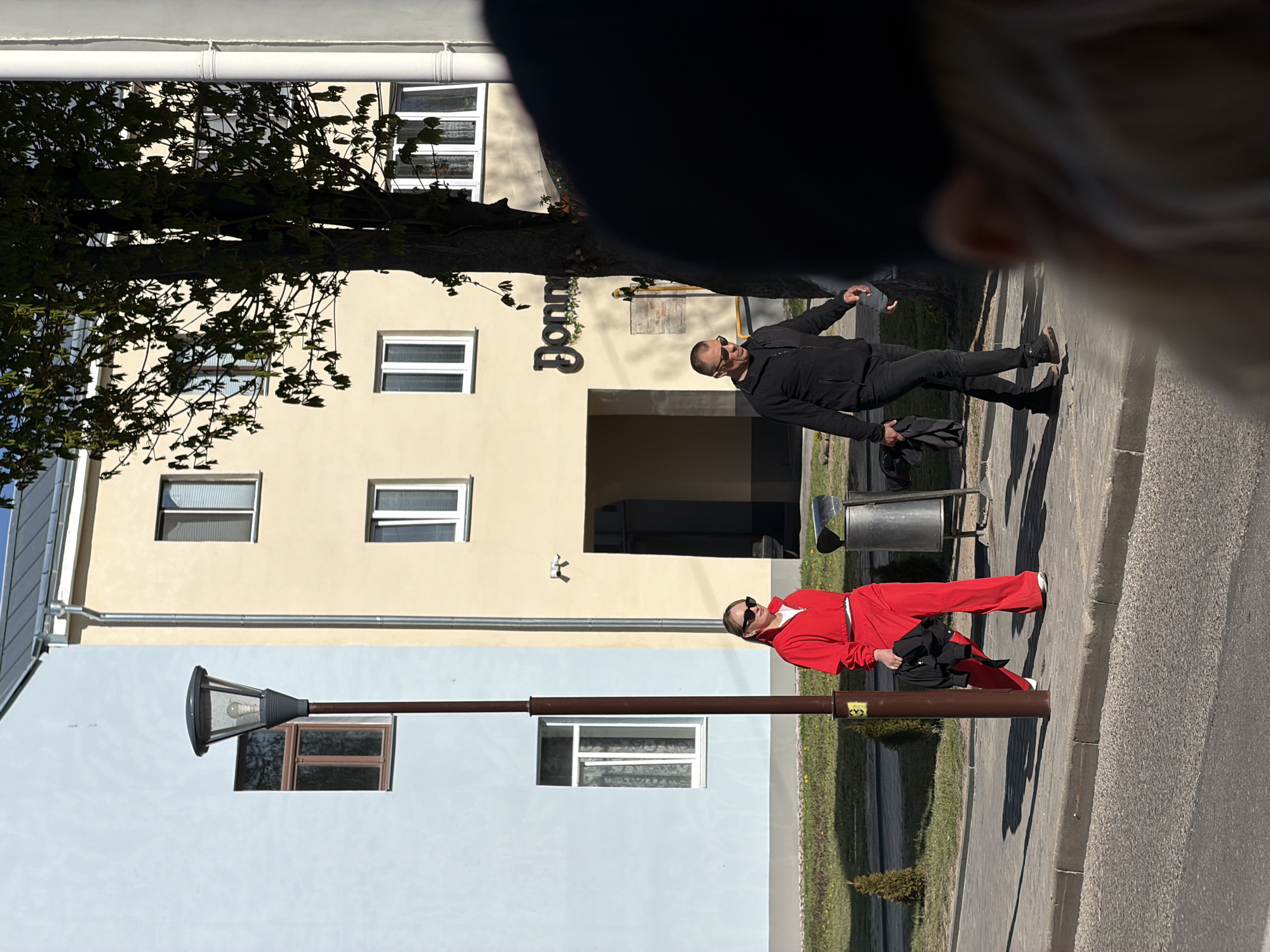 The image captures an outdoor urban scene in Hrodna, Belarus, under bright daylight. Two individuals are featured standing on a paved sidewalk in front of buildings.
The person in the foreground, positioned to the left of the frame, is wearing a distinctive bright red jumpsuit with white sneakers and dark sunglasses. They are holding a black bag or jacket in one hand and appear to be leaning casually against a dark lamppost.
Further back and to the right is a second individual, dressed entirely in black, including a jacket, pants, and shoes. They also wear dark sunglasses and hold what looks like a black item of clothing or a cap in one hand. This person stands with a relaxed posture on the sidewalk.
Behind the subjects are two main buildings: a light yellow one with several white-framed windows and a darker recessed entrance, and to its left, a light blue building with a brown-framed window and a white-framed window. A large, leafy tree partially covers the upper portion of the yellow building. The ground is paved, with areas of green grass and a small evergreen bush visible. Other street furniture includes a dark lamppost (the one the person in red is near) and a metal trash can.
The strong sunlight indicates a clear day, casting distinct shadows of the people and lamppost on the pavement. The top right corner of the image is blurred, suggesting the photo was taken from a close, slightly high perspective, possibly revealing a part of the photographer's head or shoulder.
Visible text on the light yellow building reads "Donm", likely part of a business name or sign.
