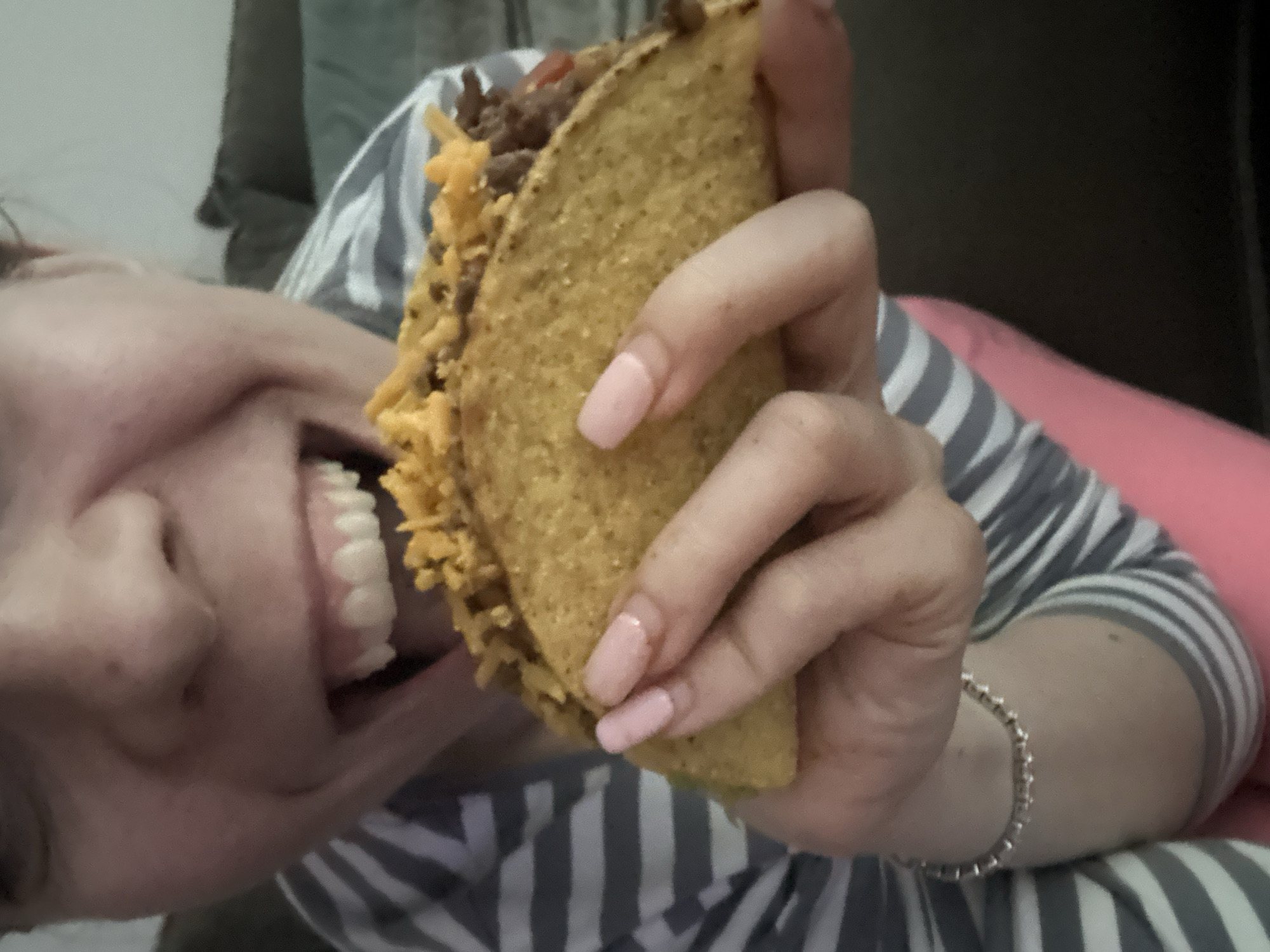 A close-up, slightly tilted image captures a person in the midst of eating a taco. The main subject is a person, likely female, whose face is seen from the nose down, turned to the left side of the frame, with a wide, joyful smile and an open mouth. She is about to bite into a hard-shell taco, which she holds firmly with her right hand.
The taco is well-filled with what appears to be seasoned ground beef and a generous amount of shredded yellow cheese spilling over the top. Hints of other toppings, possibly diced tomatoes, are also visible. The person's nails are manicured with a light pink polish. She is wearing a grey and white striped long-sleeved top, and a silver-colored chain bracelet is visible on her right wrist.
The setting appears to be indoors, with a blurry background that suggests a casual home environment, possibly a couch or bed. There are no specific location cues visible to indicate "Amsterdam, Netherlands." The lighting suggests an indoor setting, and no specific time of day can be determined. No visible text is present in the image. The overall emotion conveyed is one of pleasure and anticipation as the person prepares to enjoy the food.