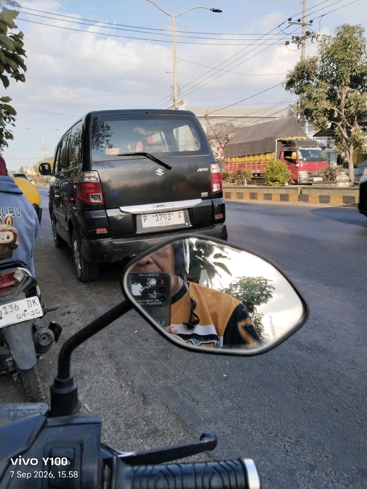 The image captures a street scene in Situbondo, Indonesia, likely on a clear day with a partially cloudy sky. In the foreground, the handlebars and part of the control panel of a motorcycle are visible, along with a side-view mirror. The mirror reflects the rider's face, wearing a yellow and black jacket, and a phone. The motorcycle's license plate, "0136 DK G9.3C", is also visible.
Behind the motorcycle, a dark-colored Suzuki APV van is parked or stopped, displaying a license plate "P 1793 F 08 26". To the left of the van, a person wearing a blue hoodie with a cartoon character on it is partially visible. Further down the street, a red truck with a covered cargo bed is seen. The road is asphalt with a yellow and black striped curb. Trees and power lines are present in the background, suggesting an urban or suburban environment. The overall scene appears to be during daylight hours, possibly late afternoon given the lighting.