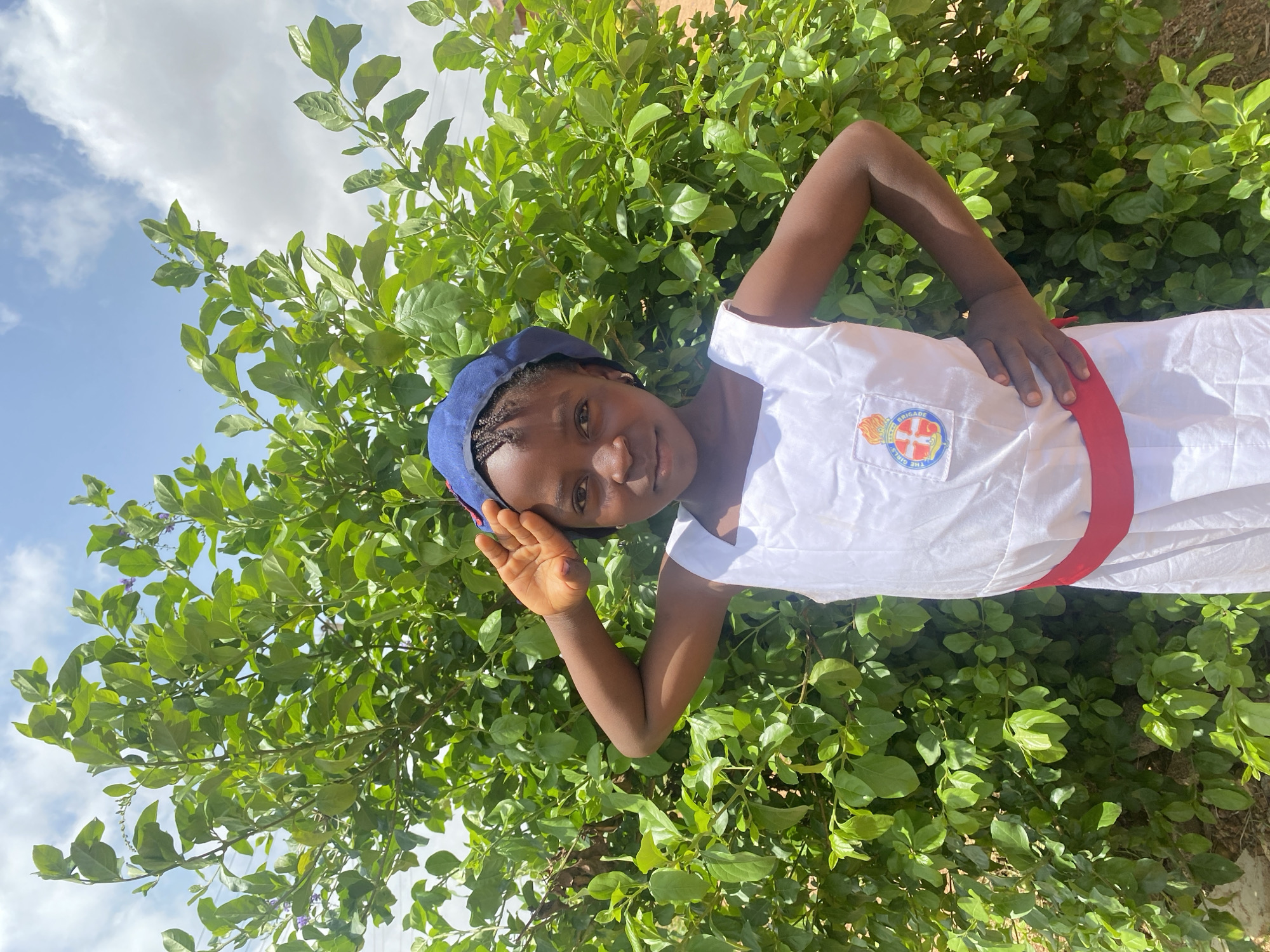 A young girl stands in front of a lush green bush under a partly cloudy sky in Turu, Nigeria. She wears a blue hat, a white uniform with a "The Girls' Brigade" logo on the chest, and a red sash around her waist. She is posing for the camera, smiling with her left hand raised.