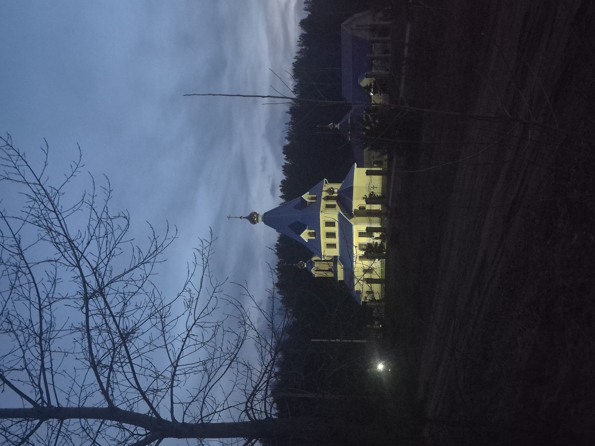 The image displays an illuminated Orthodox church or chapel at dusk or night in Chaikovsky, Russia. The central building features light-colored walls and a prominent blue, multi-tiered roof, culminating in a golden onion dome topped with an Orthodox cross. The structure is brightly lit by artificial lights, contrasting with the dark surroundings.
In the foreground, on the left side, bare tree branches are silhouetted against the twilight sky, suggesting a cold season like late autumn or winter. A dense line of dark trees, likely a forest, forms the background behind the church. The sky is a cloudy, deep blue, characteristic of twilight or early night. On the right side of the foreground, a bright street lamp casts a strong, localized light.
The scene is static, with no visible people or discernible activity. The artificial illumination of the church amidst the natural darkness creates a serene and contemplative atmosphere. No text is visible in the image.