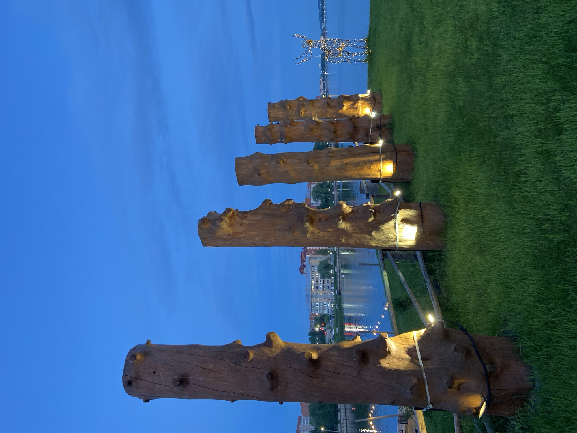 The image captures an outdoor scene at dusk, likely in Votkinsk, Russia, near a waterfront. In the foreground, a line of large, weathered wooden logs are positioned vertically on a grassy slope. These logs are illuminated from below by warm, yellow lights, creating a dramatic effect. String lights are also visible, draped along a wooden fence that separates the grassy area from the water's edge. The water itself reflects the lights from the distant shore, where buildings and trees are silhouetted against the twilight sky. To the right of the wooden logs, a decorative light sculpture of a reindeer stands prominently on the grass, its intricate outline formed by numerous small, bright lights. The sky is a deep blue, with some wispy clouds, suggesting the transition from day to night. No people are present in the immediate foreground or midground, though lights from the distant buildings suggest human activity. The overall atmosphere is serene and festive, with the artificial lighting providing a stark contrast to the natural twilight.