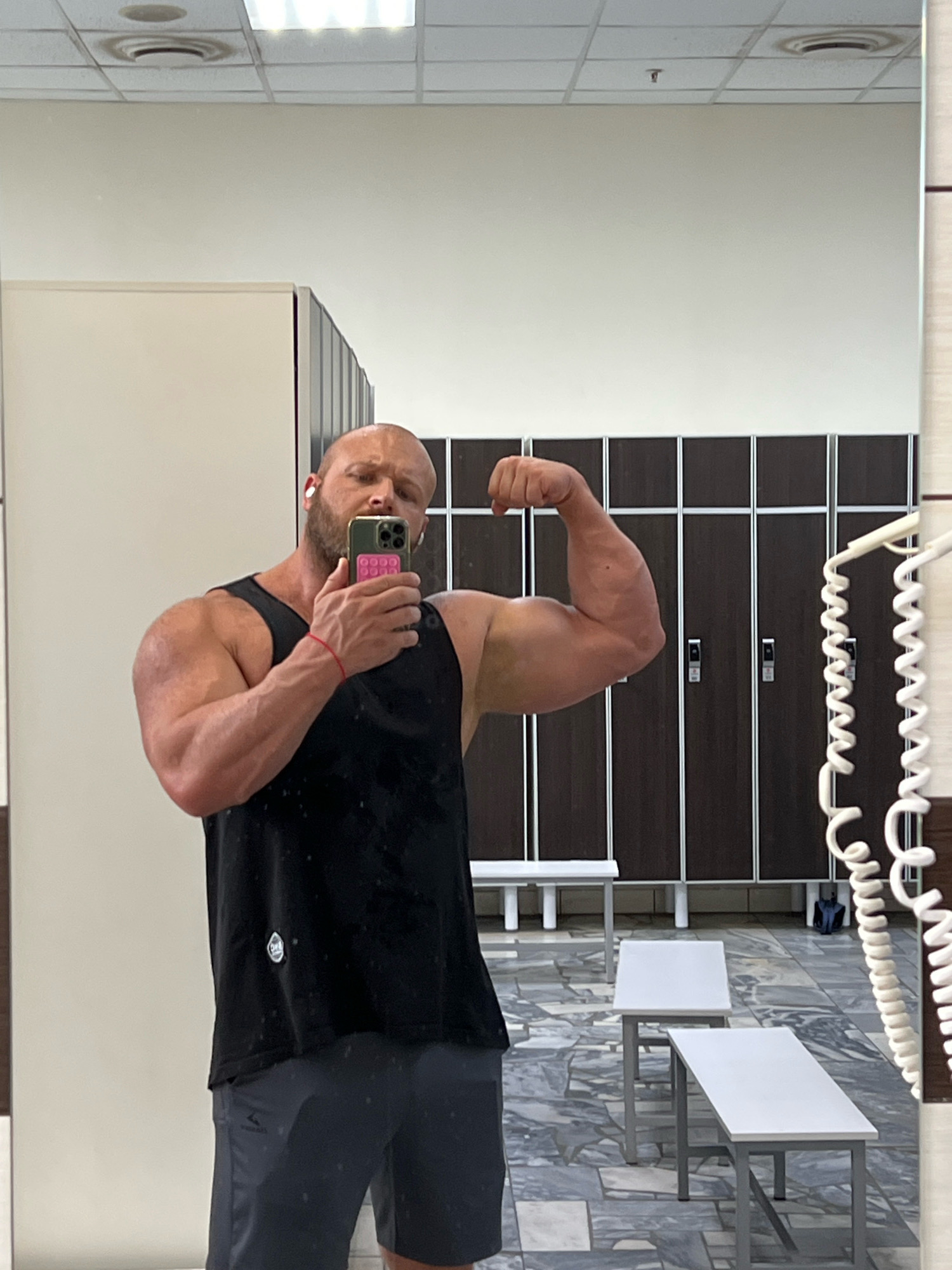 A bald, bearded man with earbuds in his ears is taking a mirror selfie in a locker room. He is wearing a black tank top and dark grey shorts. He is flexing his left bicep. In the background, there are rows of dark brown lockers and white benches. The floor is tiled with a grey and white pattern. The room appears to be well-lit, likely from overhead fluorescent lights. The presence of lockers and benches suggests this is a gym or a similar sports facility. The overall impression is that of a routine gym workout in a typical locker room environment.