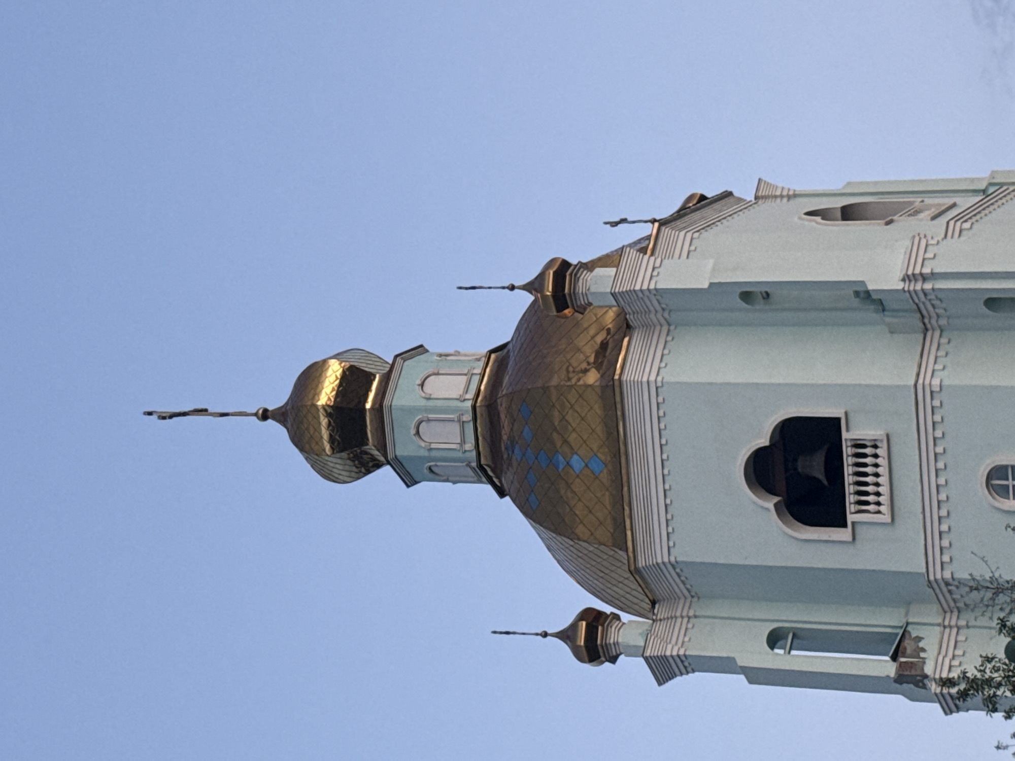 This is a close-up, eye-level, medium shot of a light blue church tower against a clear, light blue sky. The tower has several ornate golden domes topped with crosses. The largest dome is in the center of the frame, with smaller domes on either side. The tower itself is light blue with white decorative trim. There is a bell visible through an arched opening in the tower, with a white balustrade in front of it. The sun is illuminating the scene from the right, highlighting the golden domes.