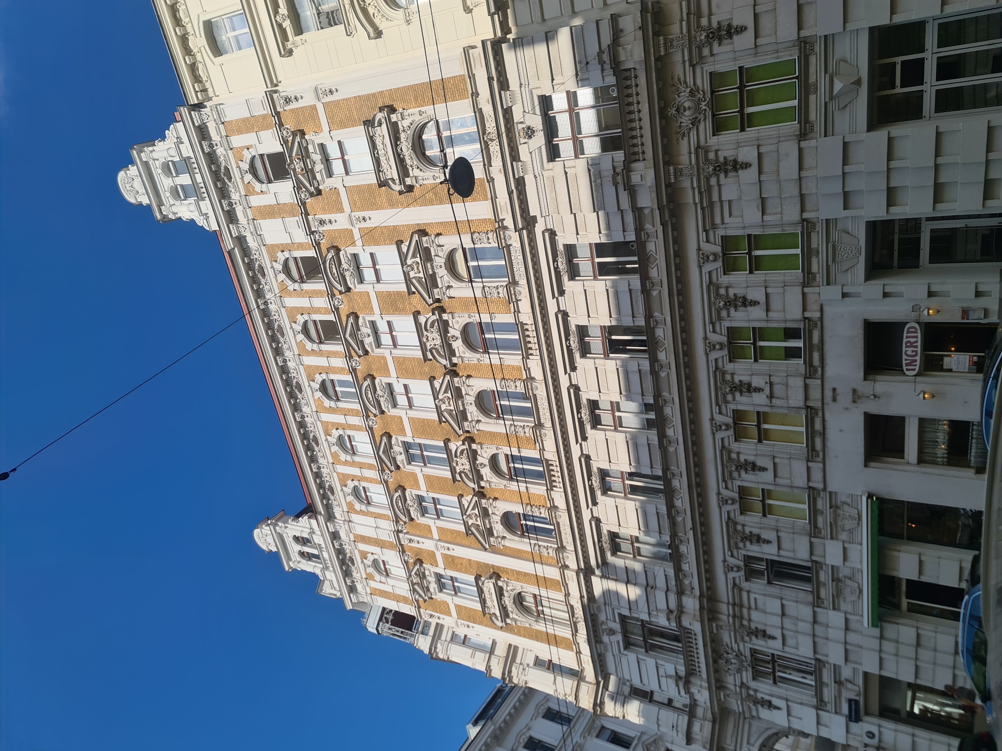 The image shows the facade of an ornate, historic building in Vienna, Austria, under a clear blue sky. The building is characterized by its yellow brickwork, white stucco decorations, and numerous windows. The architectural style appears to be Belle Époque or Secessionist, common in Vienna.
On the lower floors, there are several entrances to businesses. One prominent sign reads "INGRID" in a rounded, red and white font, indicating a business located at street level. The building's facade is detailed with intricate moldings, sculptures, and ornamental elements above and around the windows.
The perspective is from street level, looking up at the building, which suggests it is located on a city street. Utility wires are visible strung across the upper portion of the image, indicating an urban environment. Cars are partially visible in the foreground, further confirming the street-level setting. The building is brightly lit by sunlight, casting shadows on its intricate details. The overall impression is of a well-maintained, older building in a vibrant city.