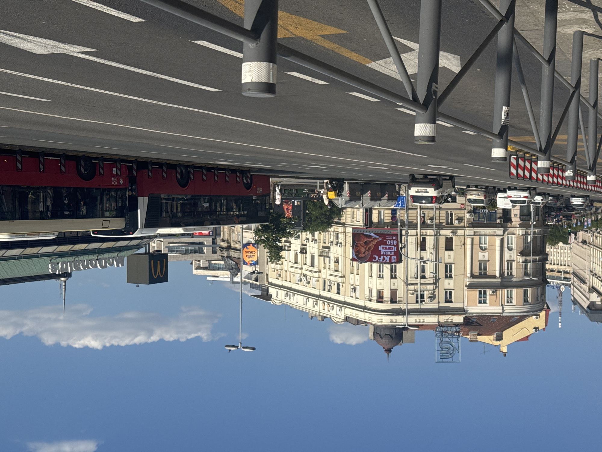 The image captures an outdoor urban scene during the daytime, under a clear blue sky with scattered white clouds. The perspective is from street level, looking up at buildings and along a road. The image is oriented sideways.
Prominent subjects include a red bus with multiple windows and a white top, parked or moving along the left side of the road. On the right side of the road, a row of cars is visible, some parked and some potentially in traffic. A metal fence with vertical posts runs along the sidewalk. Beyond the fence, several buildings with multiple stories are visible. One building has a distinctive curved facade and is adorned with many windows and architectural details. Another building features a red-tiled roof and a prominent dome.
A large McDonald's sign with its iconic "M" logo is visible on a dark rectangular structure. Further down the street, a KFC advertisement is prominently displayed on a building. Other visible text includes "Jaffa Cakes" on a yellow sign and what appears to be Serbian text on the KFC ad. The overall scene depicts a busy city street with commercial establishments. The weather appears to be sunny and pleasant.