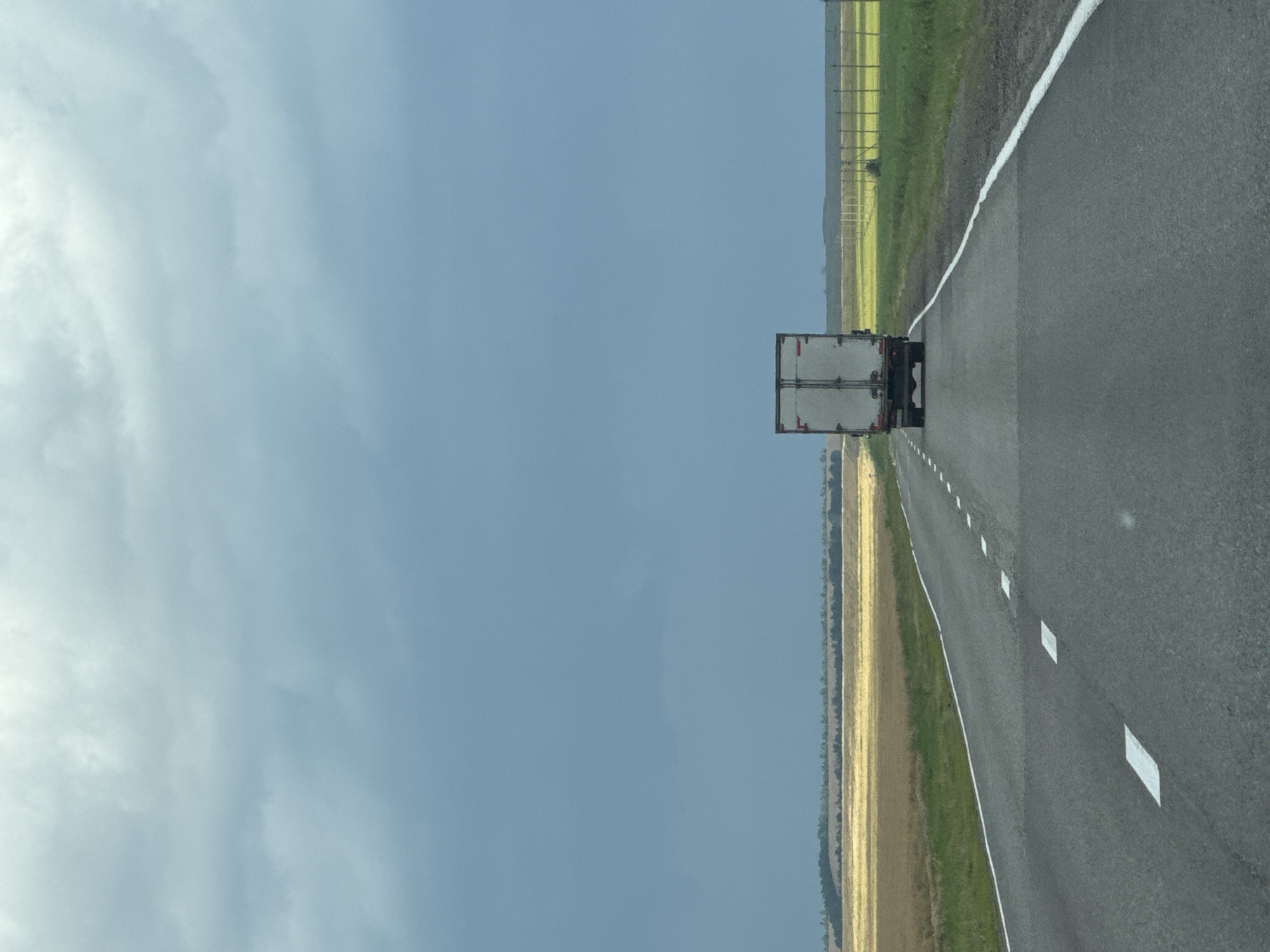 A white box truck is traveling away from the viewer on a two-lane asphalt road under a cloudy, overcast sky. The road has a white solid line on the right edge and dashed white lines marking the lanes. The landscape to the left and right of the road is agricultural, with fields of golden crops and green grass. In the distance, a treeline borders the fields. The truck is centered on the road and appears to be moving forward. The sky is a muted blue with darker clouds forming. There are no people visible in the image. The scene is one of rural travel on a highway.