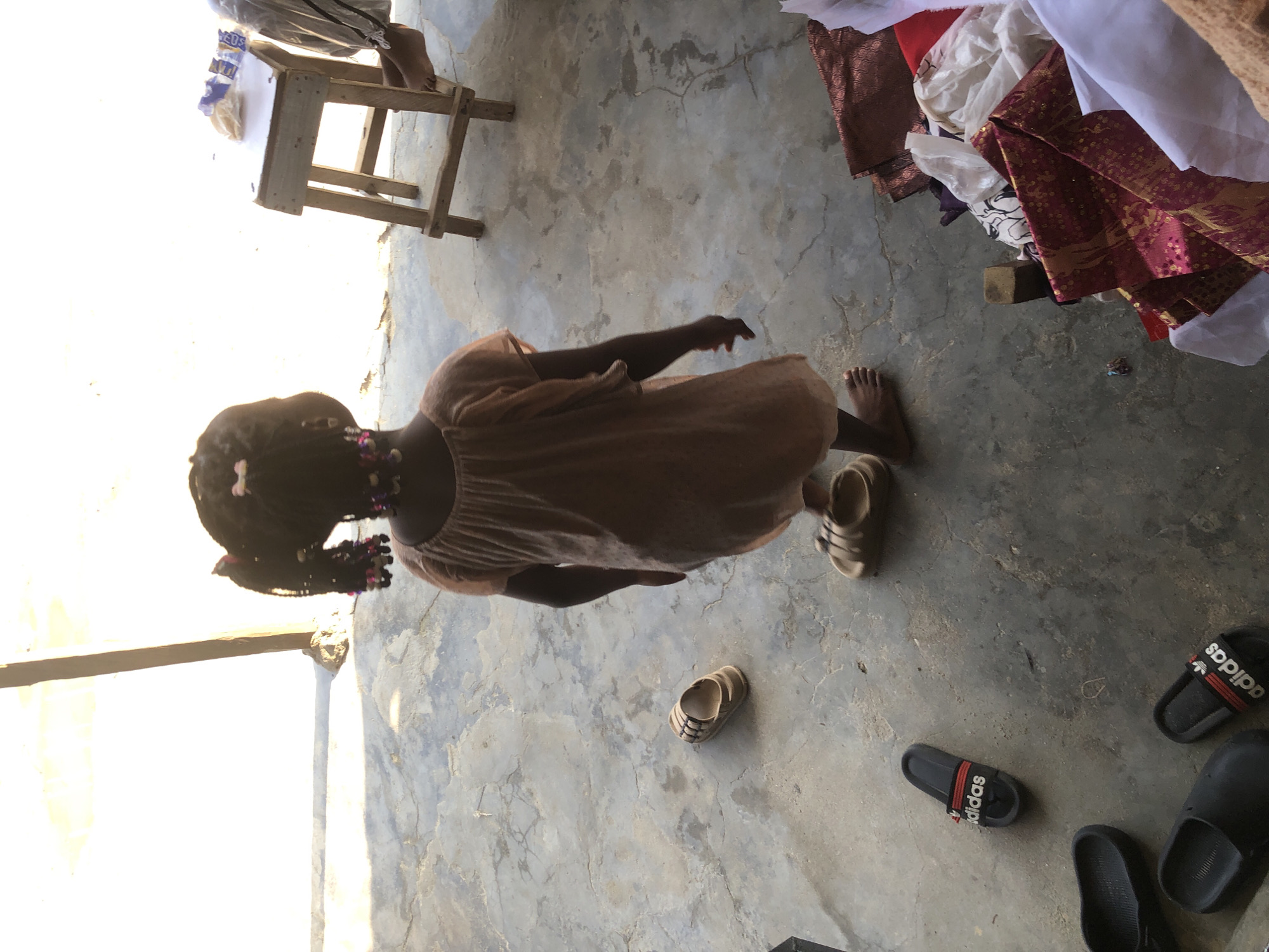 A young Black girl, viewed from behind, stands on a concrete surface. She has dark hair braided with colorful beads and wears a light orange dress. Her feet are bare, and she appears to be standing on a pair of tan sandals. Around her are several other pairs of footwear, including black Adidas slides and black clogs. To the right, a small wooden stool with a white surface holds a few items, and a person's feet are visible beneath it. In the foreground and to the right, a pile of colorful fabrics, including a richly patterned burgundy and gold textile, is present. The environment suggests an outdoor or semi-enclosed area, possibly in Ado, Nigeria, characterized by concrete flooring and natural lighting.