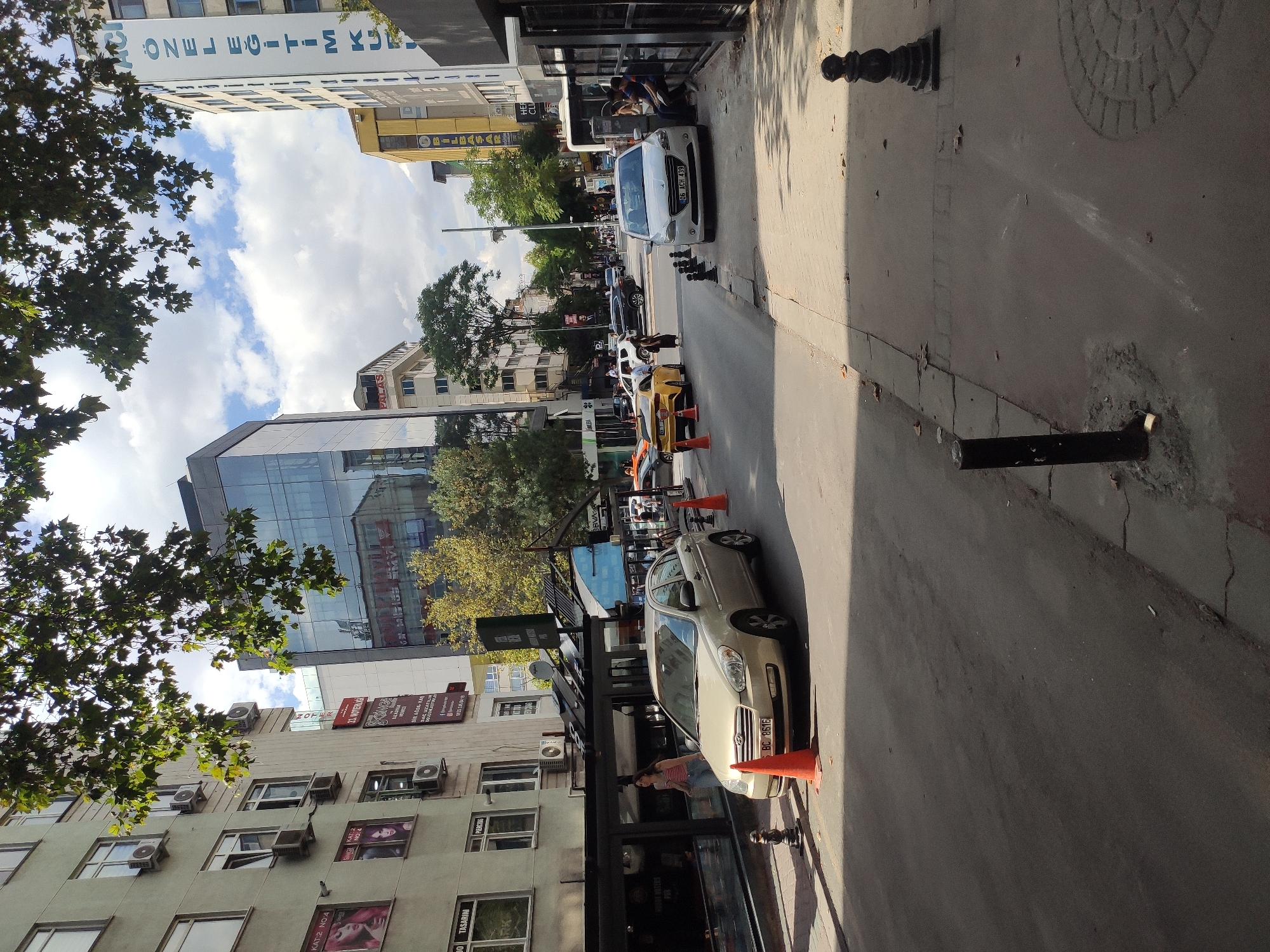 This image captures a street scene in the city of Ankara, Turkey, likely during daytime given the bright sunlight and clear sky with scattered clouds. The perspective is from a street level, looking down a road lined with buildings on both sides.
On the left, several multi-story buildings are visible. The closest one has an exterior clad in what appears to be light-colored panels or concrete, featuring numerous windows and air conditioning units. Advertisements, featuring images of women, are affixed to the facade. Some of these advertisements have text in Turkish, such as "KAT 2 NO:4" and "TASARIM," suggesting commercial establishments or services within. Below these buildings, a dark-colored awning or roof extends over a storefront, with a woman in a striped top and jeans walking near it.
The street itself is paved asphalt and appears to be a two-way road with parked cars and traffic. A light-colored sedan is parked in the foreground on the right side of the road, with a license plate visible. In the street, there are several orange traffic cones, indicating some form of traffic management or warning. Further down the street, other vehicles are visible, including a yellow sports car and a white sedan.
On the right side of the street, there are more buildings. One prominent building has large vertical text in Turkish, potentially advertising a service or institution. Another building features a modern glass facade. People are seen sitting on a low wall or ledge in front of one of the buildings on the right.
The environment includes trees, with branches and leaves overhead partially obscuring the sky in the upper part of the frame. The overall impression is of an urban environment with commercial activity and everyday street life.