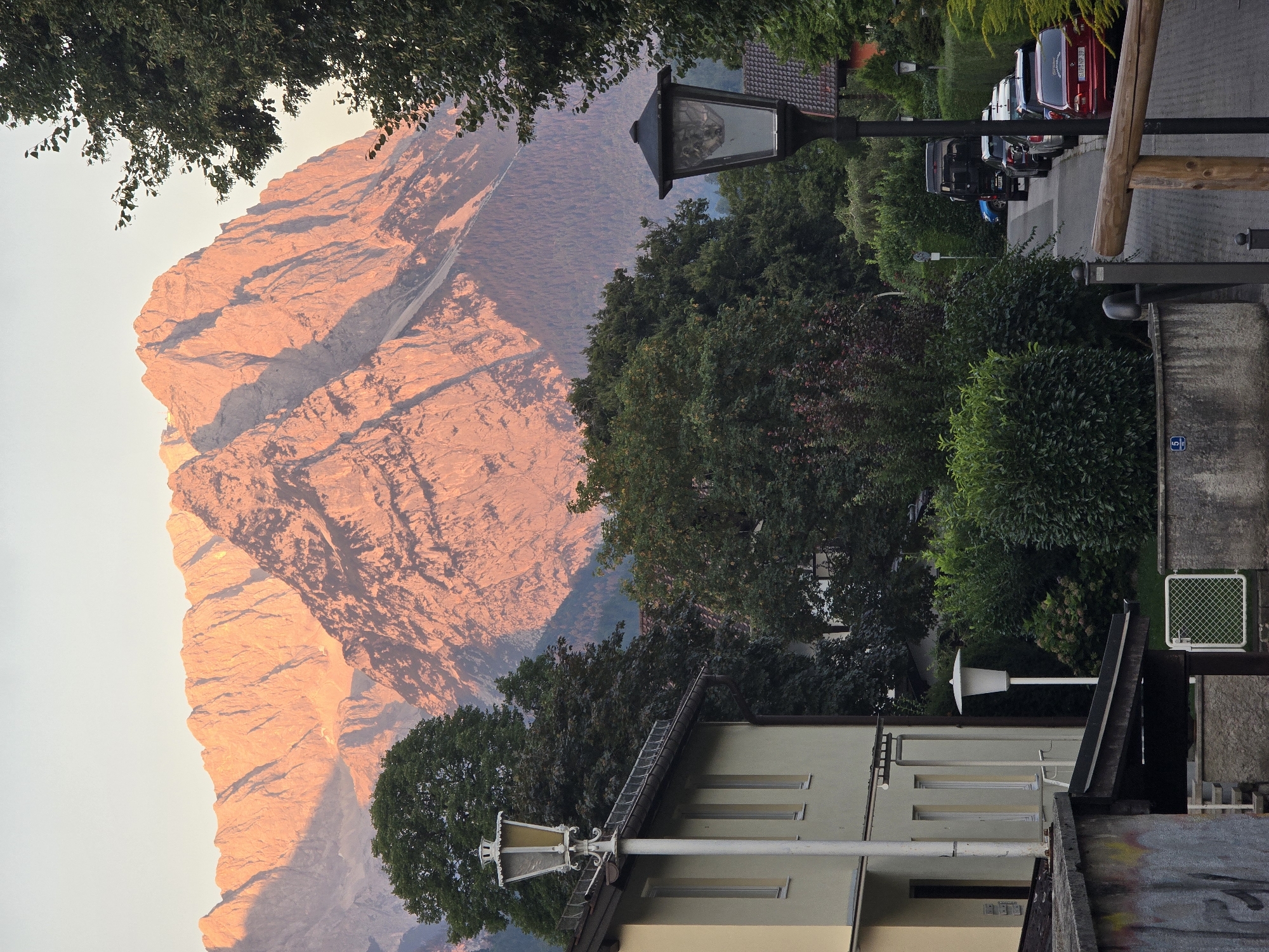The image depicts a street scene in Garmisch-Partenkirchen, Germany, with a dramatic mountain range illuminated by the setting sun in the background. In the foreground, a streetlamp with a black metal frame stands on the right side of a paved road. To the left, a portion of a cream-colored building is visible, featuring windows and a roofline. Further down the street, several parked cars are lined up, including a red car with a German license plate, a black van, and other vehicles. Lush green trees and bushes are abundant throughout the scene, framing the street and the building. The sky above is a clear, pale hue, indicative of early evening. The mountain range, in the distance, displays sharp peaks and deep shadows, with the warm, rosy light of the sunset casting an ethereal glow on its rugged surfaces.