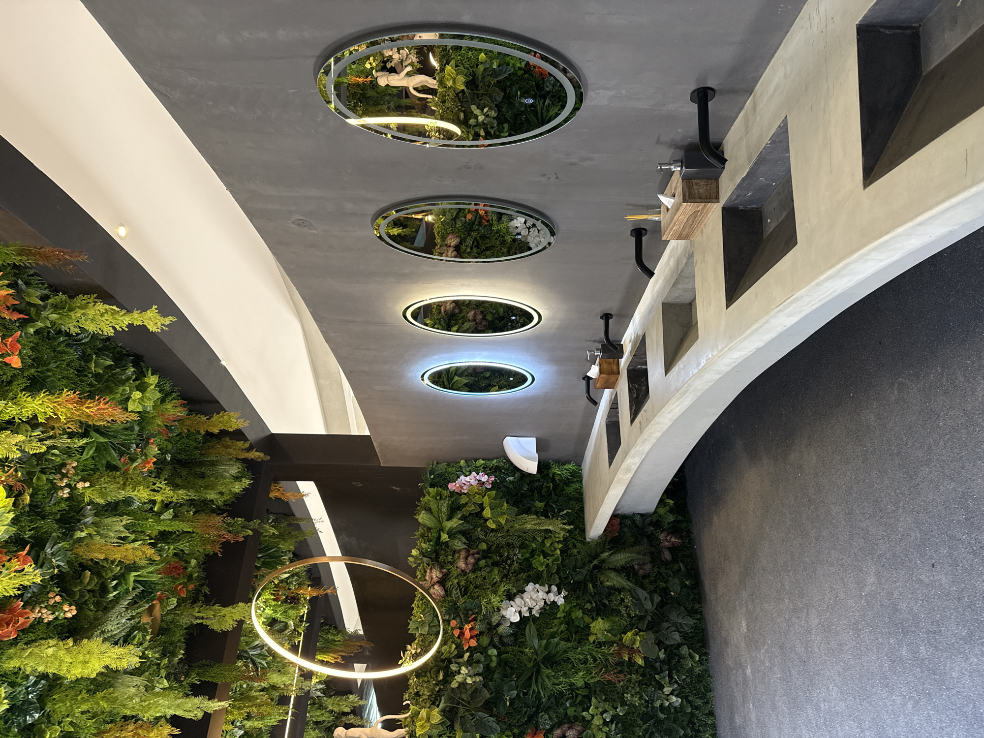 The image depicts a modern restroom with a jungle-themed decor in Ko Samui District, Thailand. The restroom features a long, curved concrete countertop with multiple sinks and black faucets. Above the sinks, a wall is covered with an abundance of artificial greenery, including hanging vines and flowering plants, creating a lush, botanical atmosphere. To the left, a circular light fixture hangs from the ceiling, surrounded by more hanging artificial plants. Three oval mirrors with integrated LED lighting are mounted on the gray concrete wall above the countertop. Each mirror reflects the lush greenery and a white monkey statue. The floor is a dark gray, textured surface. A white automatic hand dryer is mounted on the green wall between the sinks and the mirrors. The overall impression is a contemporary and nature-inspired design for a public restroom.