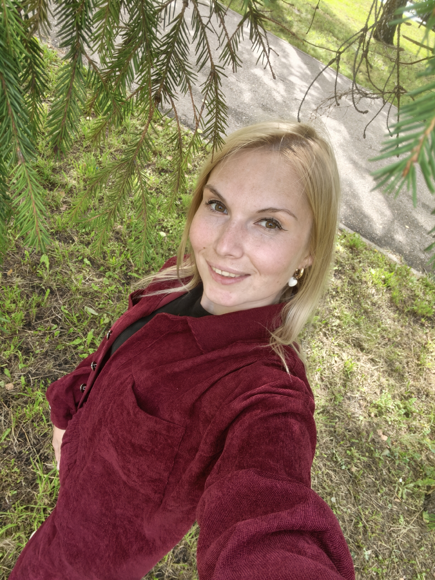 A blonde woman with freckles smiles at the camera, taking a selfie under the branches of a fir tree. She is wearing a burgundy corduroy jacket over a black shirt and gold hoop earrings with a white heart-shaped charm. The ground around her is covered in green grass and dry leaves, with a paved path visible in the background. The scene appears to be in a park or outdoor area within the city of Sterlitamak, Russia.