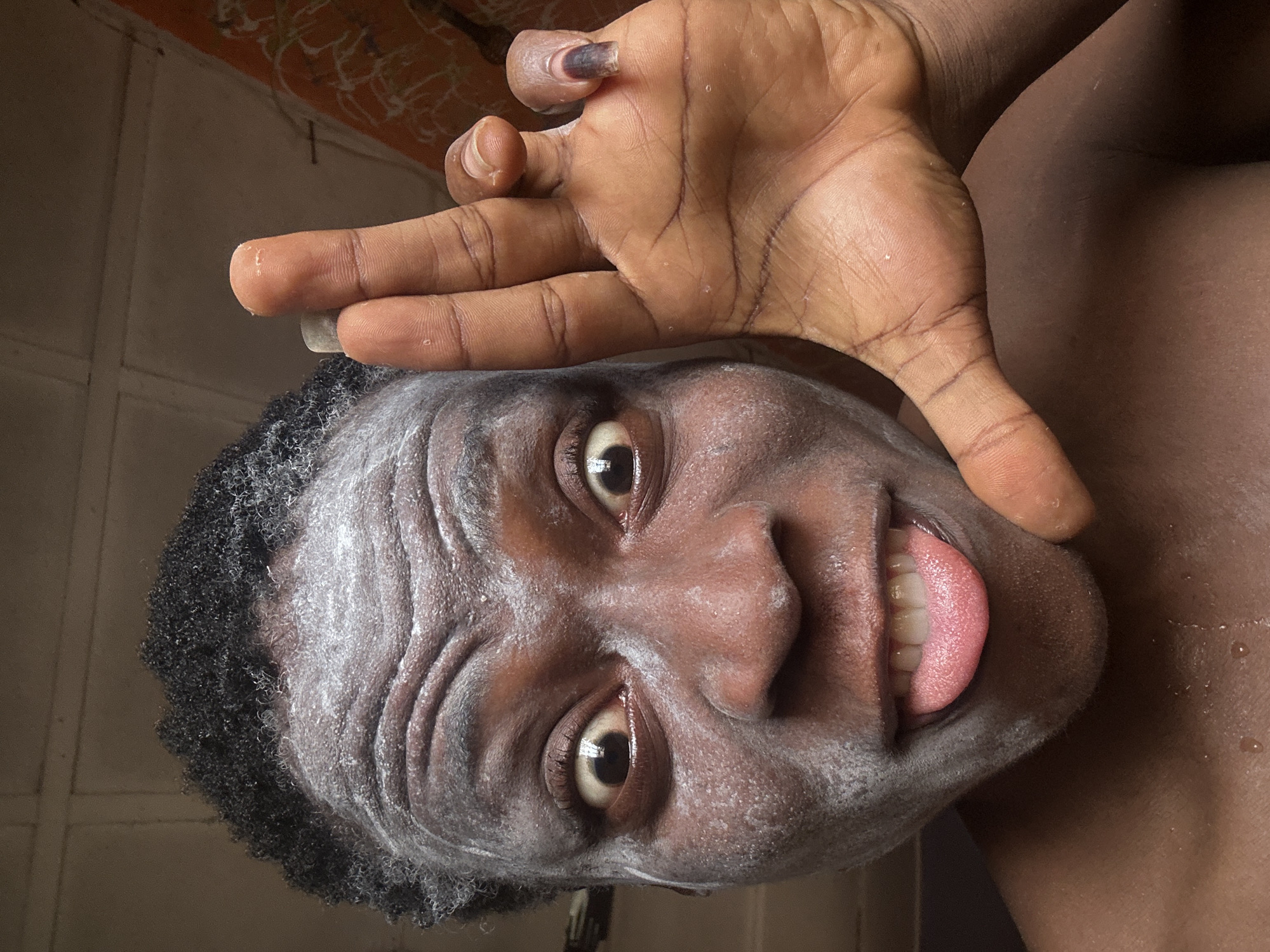 A close-up, tilted view shows a Black person with short, curly hair and a white substance covering their face, possibly a cosmetic mask. They are sticking their tongue out and making a playful expression. Their right hand is raised, with fingers spread. The background is a blurred wall, and the location is Unknown, Unknown.