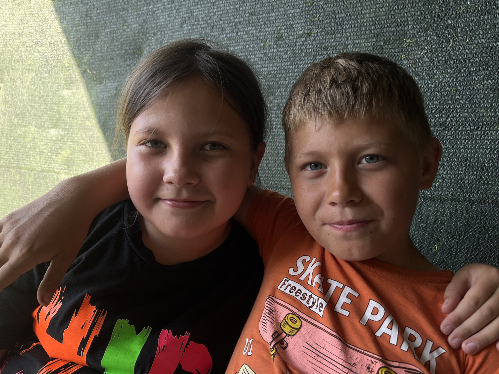 This media file is a close-up, medium shot portrait of two young children, a girl and a boy, seated closely together. The girl is on the left, with shoulder-length brown hair, looking directly at the camera with a slight smile. She is wearing a black t-shirt with colorful, abstract brushstroke designs in orange, green, and pink. The boy is on the right, with short blonde hair and blue eyes, also looking at the camera with a subtle smile. He is wearing an orange t-shirt with a graphic that includes the text "SKATE Freestyle" and an image of a skateboard.
The children are positioned with their shoulders touching, and the girl's arm is around the boy's shoulders, indicating a close relationship, possibly siblings. Their faces are well-lit, suggesting it's daytime. The background is a textured, dark green mesh material, with a diagonal band of lighter green and yellow light on the left side, possibly indicating sunlight filtering through a screen or awning. The lighting and the children's relaxed expressions suggest a casual, outdoor setting. No specific time of day or weather conditions are definitively evident, but the lighting points to daylight. The location context provided is Novopokrovskaya, Russia, but there are no explicit visual cues in the image to confirm this. The overall mood is warm and familial.