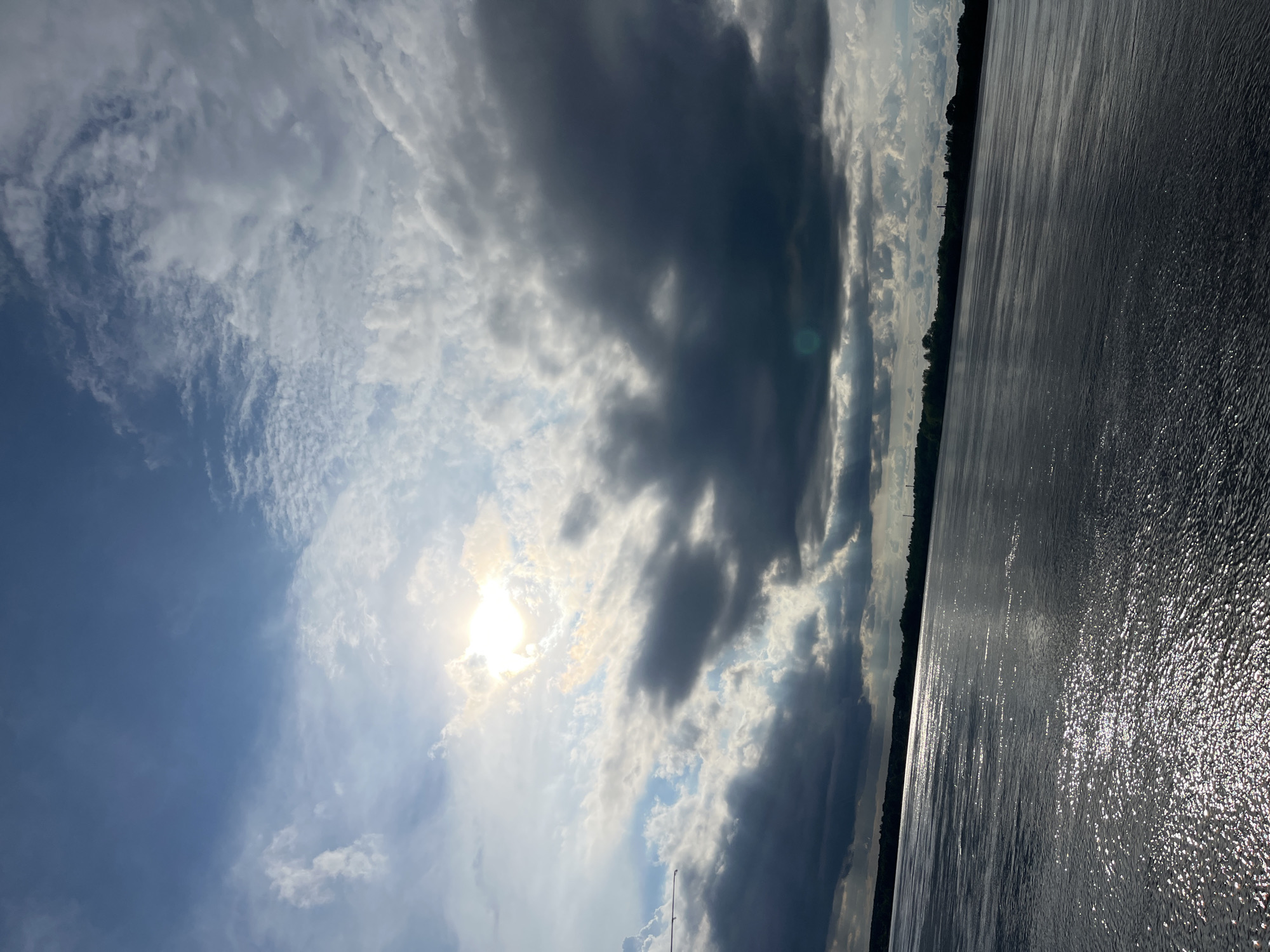 The image shows a body of water, possibly a lake or a wide river, with a tree-lined shore in the distance. The sky is filled with dramatic clouds, ranging from bright white to dark gray, with the sun partially obscured. Sunlight reflects brightly off the water's surface. A fishing rod is barely visible near the bottom center of the frame.
Given the prompt's request for the location in Chaikovsky, Russia, and considering the characteristics of water bodies in that region, the location could be on the shore of the Kama Reservoir near Chaikovsky, Perm Krai, Russia.