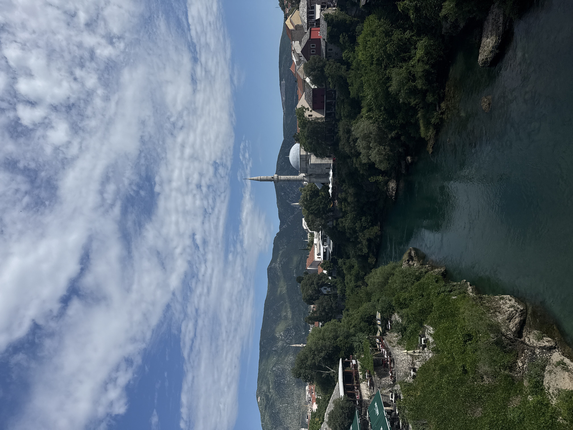 The image presents a wide-angle view of the Neretva River flowing through the city of Mostar, Bosnia and Herzegovina, bordered by steep, vegetated banks and backed by mountains under a partly cloudy sky.
The upper third of the image shows a bright blue sky partially covered by scattered white cumulus and stratocumulus clouds. Below the sky, in the background, a range of green, rugged mountains with visible rock formations spans the horizon.
In the midground, the city of Mostar is visible along the riverbanks. On the left side, multiple levels of terraced outdoor cafes, identifiable by dark green awnings (some marked "KABALAK"), are built into the hillside, surrounded by dense green trees and shrubs. Tables and chairs are set up, suggesting active establishments. Further into the distance on the left, a slender white minaret can be partially seen rising above the trees.
On the right side of the river, a prominent stone mosque featuring a large grey dome and a tall, slender, pointed minaret dominates the skyline (likely the Karagöz Bey Mosque). Adjacent to the mosque, a cluster of traditional buildings with red-tiled roofs and light-colored facades, some with red or yellow walls, extends along the riverbank, nestled among abundant green foliage.
The foreground is dominated by the Neretva River, which flows from the distant city towards the viewer. The river water is a distinctive deep emerald green or turquoise hue. The banks are steep, rocky, and heavily covered with lush green trees and bushes, providing a natural setting for the urban landscape. No individual people or explicit actions are clearly discernible due to the distance.