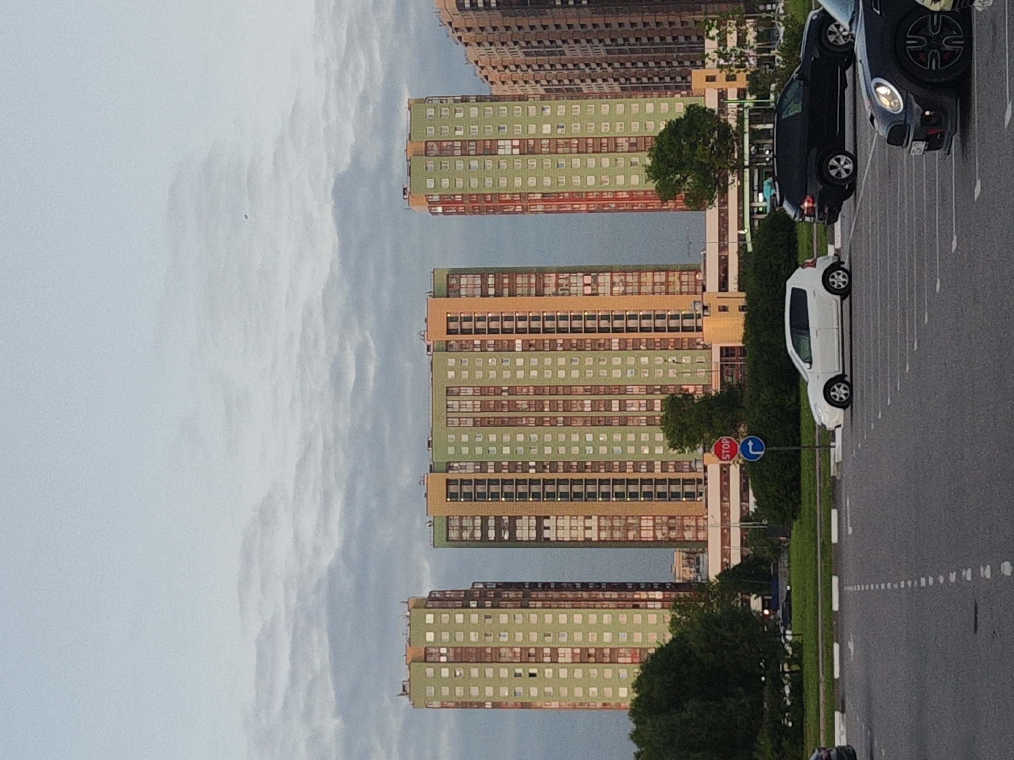 This image captures an urban or suburban residential scene, likely in Saint Petersburg, Russia, based on the architectural style. The photo appears to be taken from a slightly elevated position, looking across a parking area towards a series of tall apartment buildings under an overcast sky.
The prominent subjects are several multi-story apartment blocks lining the right side of the frame. These buildings share a distinct design, characterized by light green facades punctuated by horizontal bands of orange-brown, possibly indicating balcony or window sections. Their uniform, modular appearance is typical of residential developments in the region.
In the foreground and midground on the left, a paved parking lot or road features multiple vehicles. A dark-colored Mini Cooper or similar compact car is partially visible in the immediate foreground, followed by a black SUV or crossover, and a white compact car further down the road. White painted lines mark parking spaces or lanes on the asphalt.
Between the road and the buildings, a strip of green grass is visible, along with several trees and shrubs, adding natural elements to the urban landscape.
The sky, occupying the upper portion of the frame, is entirely overcast with soft, light gray clouds, diffusing the daylight. A tiny, dark speck, possibly a bird, can be observed in the upper sky.
Two traffic signs are visible in the midground: a red octagonal sign bearing the word "STOP," and a blue circular sign with a white arrow indicating a mandatory right turn.
The scene appears calm and devoid of overt human activity, with parked cars suggesting a quiet moment during the day. The diffused light suggests it could be morning, late afternoon, or simply a heavily cloudy day.