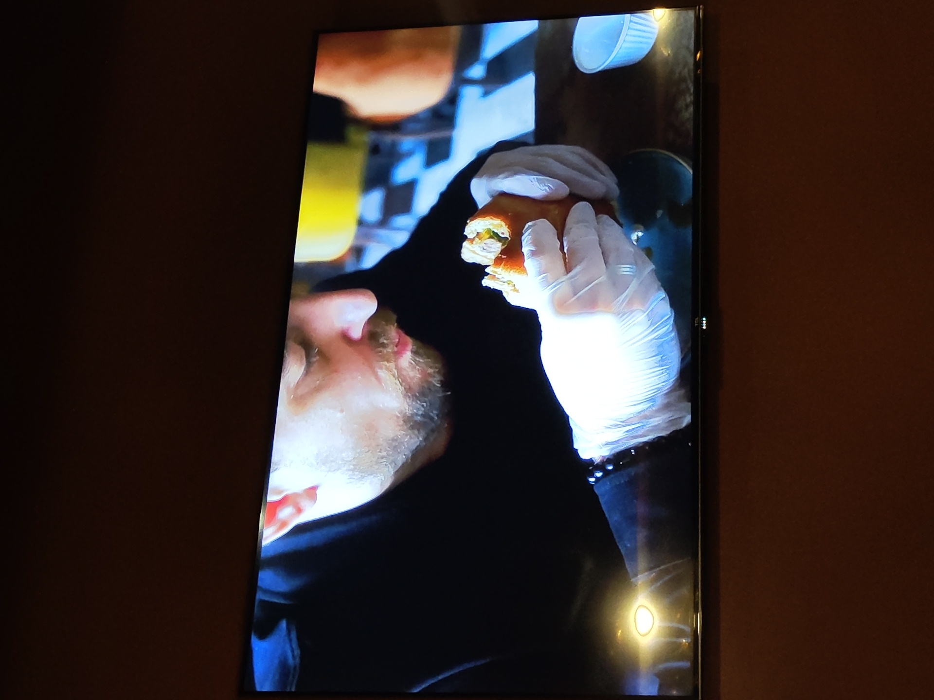 A medium shot captures a man wearing disposable gloves and a black hoodie, holding a partially eaten sandwich. The man has short, light brown hair, a beard, and appears to be in his late 30s or early 40s. He is seated at a table with a dark wooden surface. To his right, a small white paper cup is visible. In the background, a blurred checkered floor and yellow and orange seating can be seen, suggesting an indoor dining environment, possibly a cafe or restaurant. The image is a photograph of a television screen. The man is looking down at the sandwich. The television is mounted on a dark brown wall.