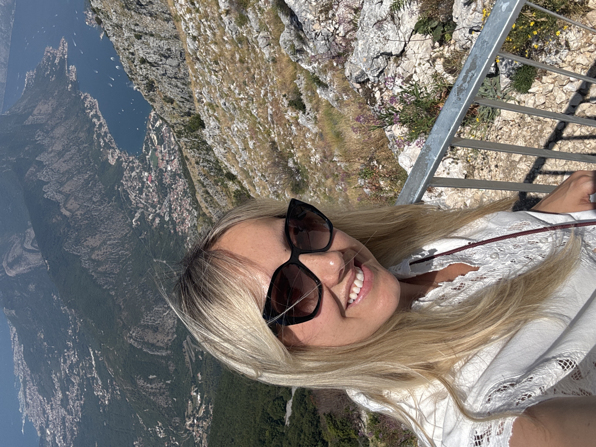An outdoor, daytime, selfie shows a young woman with blonde hair and sunglasses smiling at the camera. She is wearing a white lace-trimmed blouse. She is standing on a rocky terrain with a railing in the foreground. In the background, a body of water is visible, with a city nestled along its shore and surrounded by mountains. This location is identified as the Bay of Kotor, near Kuk, Montenegro. The Bay of Kotor is a UNESCO World Heritage site known for its dramatic fjord-like appearance and historic towns.