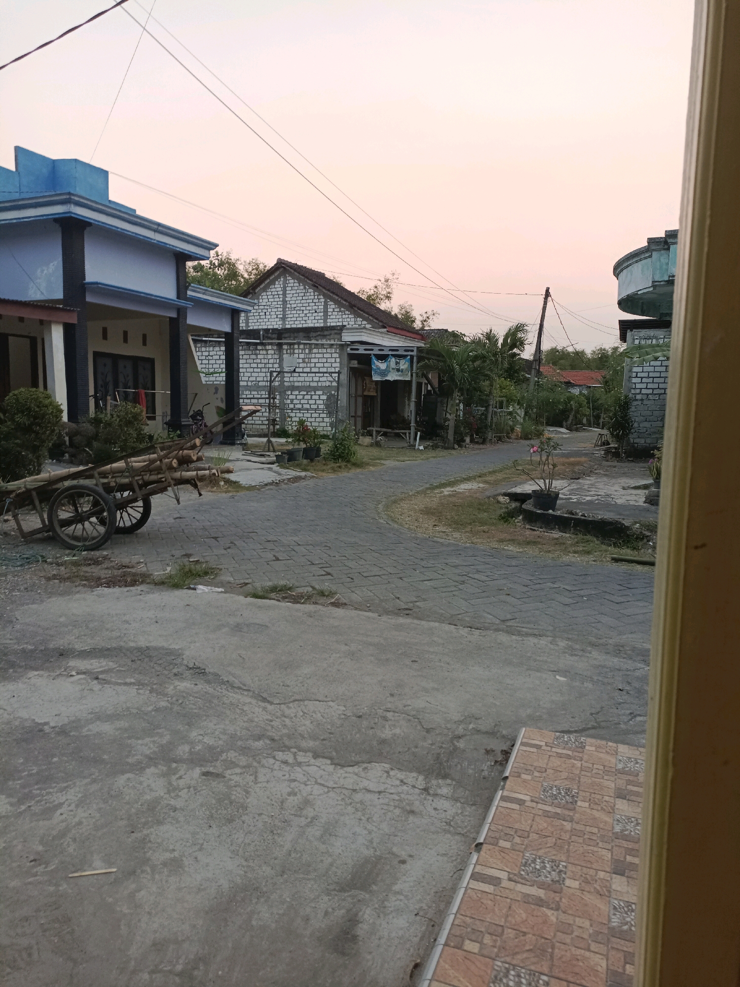 The image depicts a street scene in Rancang Kencono, Indonesia, at dusk or dawn. A paved road with interlocking brick patterns curves through a residential area. On the left, a light blue and white two-story house with dark pillars stands behind a small patch of grass and bushes. In front of the house, a wooden cart laden with logs is parked on the side of the road. Further down the street, more houses are visible, some constructed with white brick. Trees and foliage are scattered throughout the scene, and overhead power lines crisscross the sky, which is a soft gradient of pink and yellow, indicating twilight. The overall impression is a quiet, rural or semi-urban neighborhood.