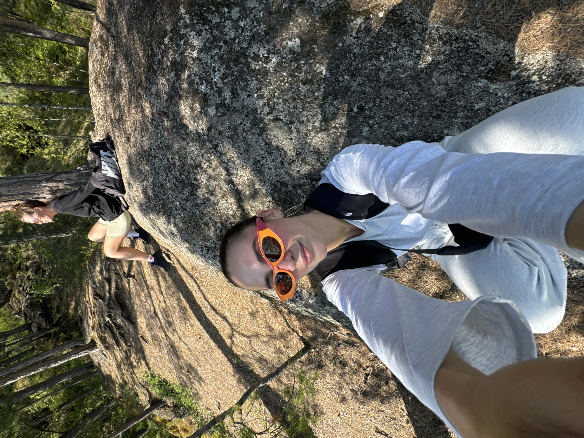 The photograph appears to be a selfie taken outdoors. The person taking the photograph has orange sunglasses and is wearing light-colored clothing. Another person is visible in the background, wearing a dark shirt and light shorts and standing near a large rock. The environment includes trees, sunlight, and what appears to be a wooded area. Based on the location information provided, this is likely a natural area near the city of Krasnoyarsk, Russia.