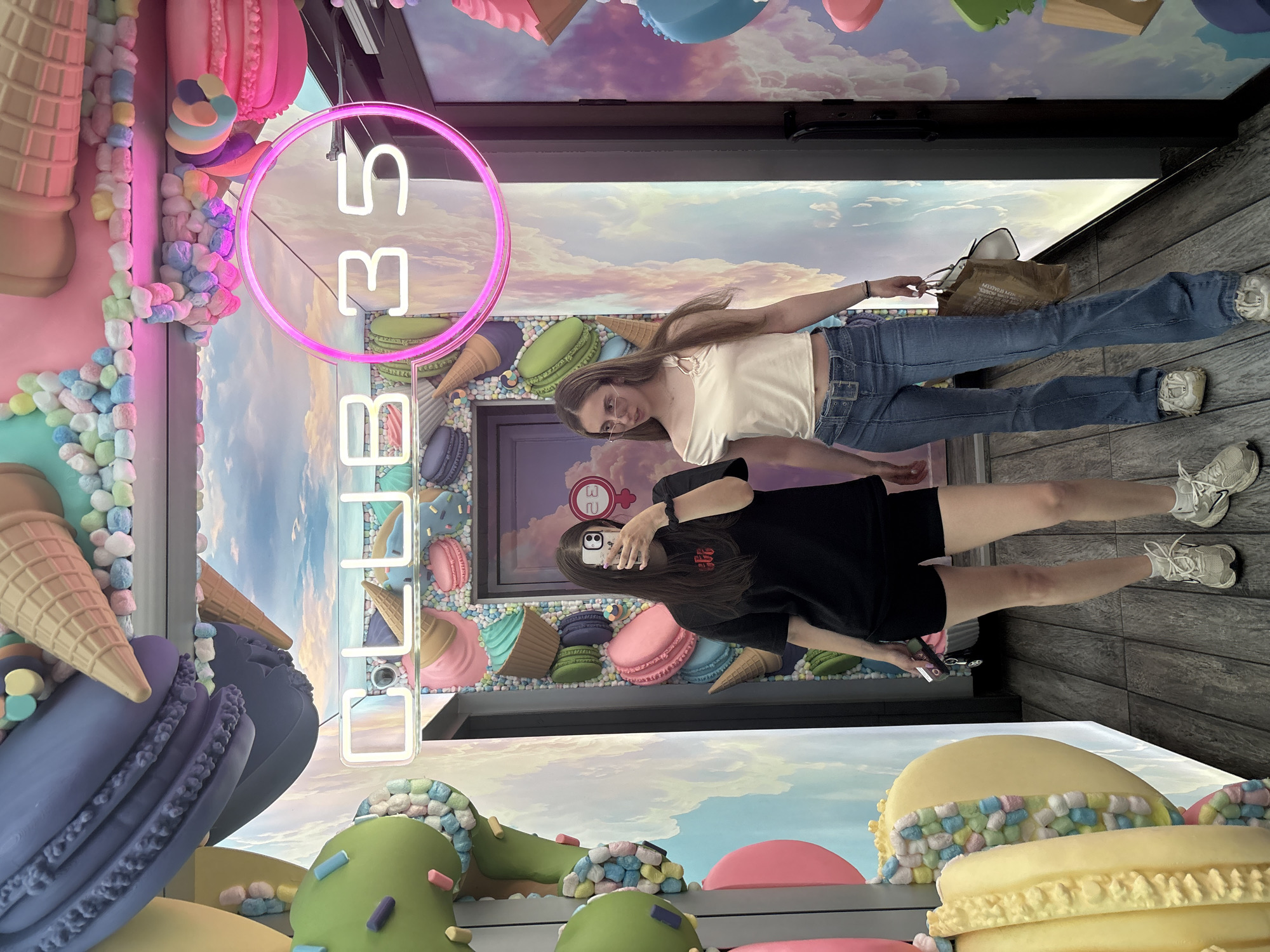 Two young women are posing for a selfie in an indoor amusement park or attraction decorated with oversized candy and dessert replicas. The scene is vibrant and colorful, with a sky-like backdrop and a neon sign displaying "CLUB 35". The woman on the left wears a black t-shirt and black shorts, while the woman on the right is dressed in a white off-the-shoulder top and blue flared jeans. The woman on the left holds a phone and keys, while the woman on the right carries a paper bag. The floor is made of wooden planks. The location is identified as Sofia, Bulgaria.