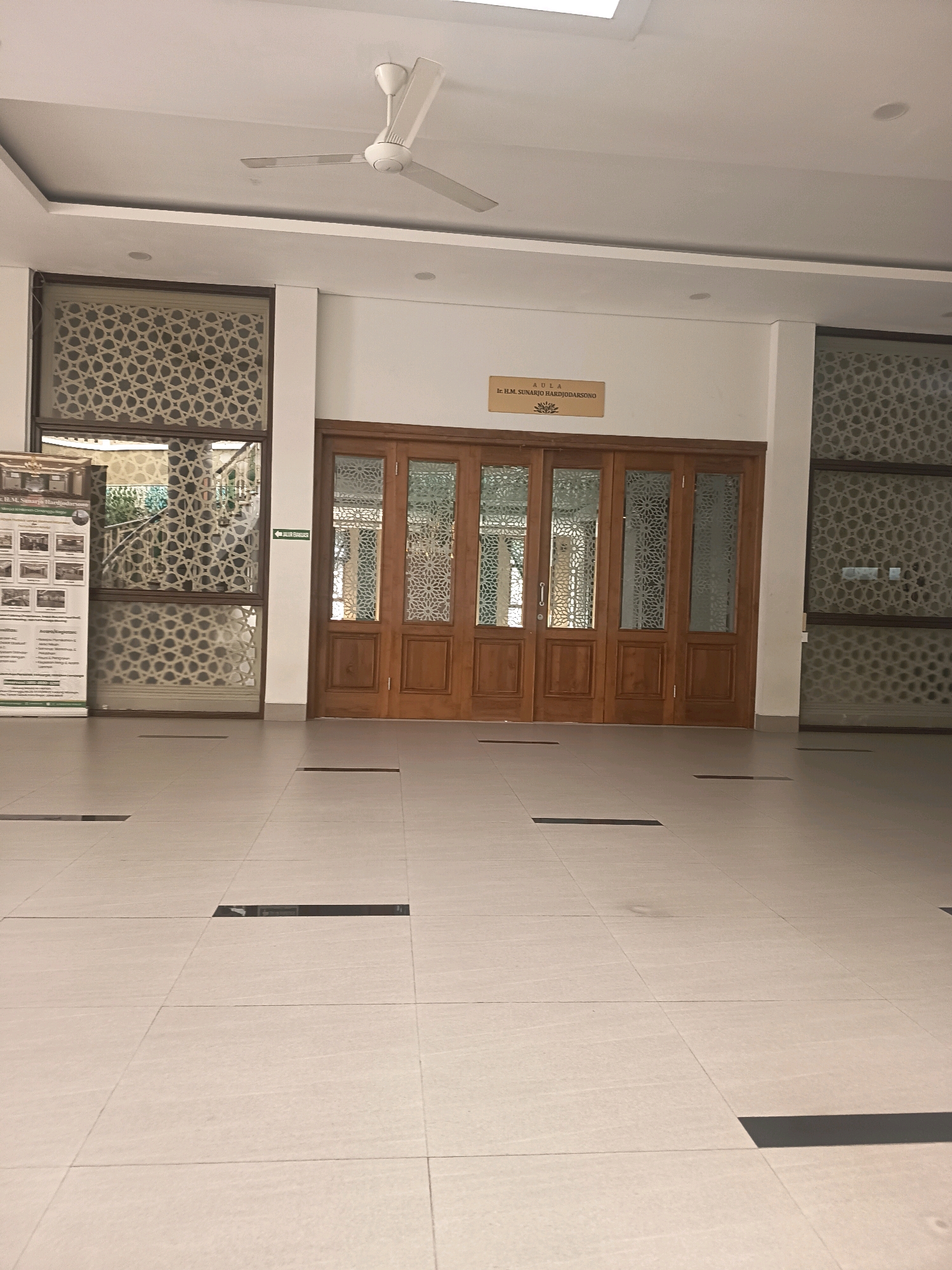 The image shows the interior of a building, likely a public space, with a tiled floor and a ceiling fan. In the background, there are large wooden doors with glass panels featuring intricate geometric patterns. To the left of the doors, there is a large window with similar decorative glass and a banner on display. Above the doors, a plaque reads "AULA Ir. H.M. SUNARJO HARDJODARSONO". The overall impression is one of a well-maintained and possibly formal or institutional environment. This location is identified as the Aula Ir. H.M. Sunarjo Hardjodarsono building at the IPB University, in Bogor, Indonesia. The scene is devoid of people.
