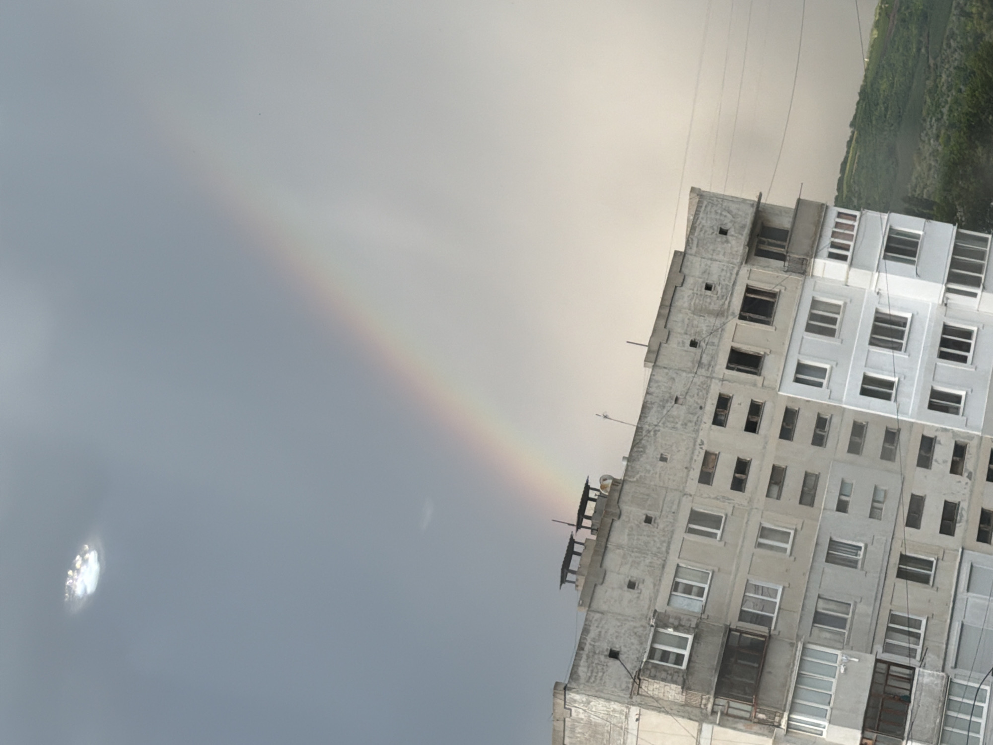 The image captures a view of an apartment building in Strășeni, Moldova, under a cloudy sky featuring a faint rainbow. The building is a multi-story, concrete structure with numerous windows and balconies. The architecture suggests a Soviet-era design. The sky is overcast, with a bright, circular cloud formation visible in the upper left corner. A subtle rainbow arcs across the upper portion of the frame, extending from the upper left towards the right. The lower right of the image shows a sloping, green hillside covered in trees, indicating a semi-rural or suburban setting within the city. No people are visible in the image. The overall scene is one of a slightly gloomy day with a natural phenomenon adding a touch of color.