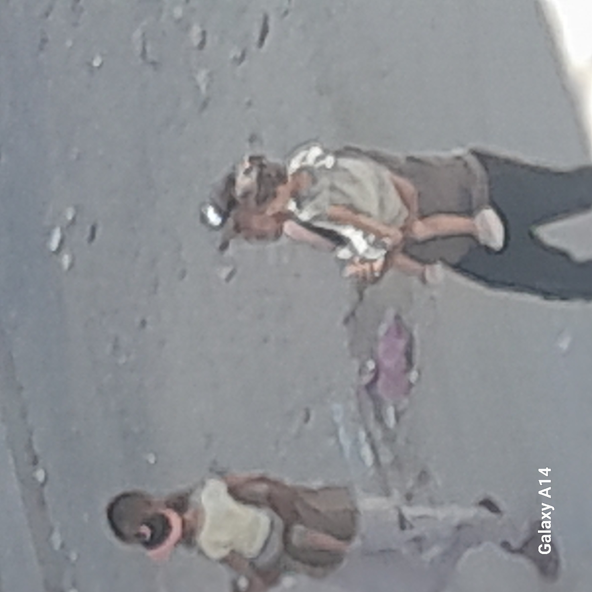 In Khan Yunis, Palestinian Territories, two individuals, likely a parent and child, are walking on a dirt or gravel path. The individual in the foreground, appearing to be an adult, is carrying a young child. The adult is wearing dark pants and a dark shirt, and the child is wearing a light-colored shirt with a patterned design and a light-colored hat. To the left, a smaller child wearing a pink hat and a light-colored shirt is also visible, possibly walking ahead or to the side of the adult. The background consists of a textured, sandy or gravelly ground with some scattered debris. The lighting suggests an outdoor, daytime scene.