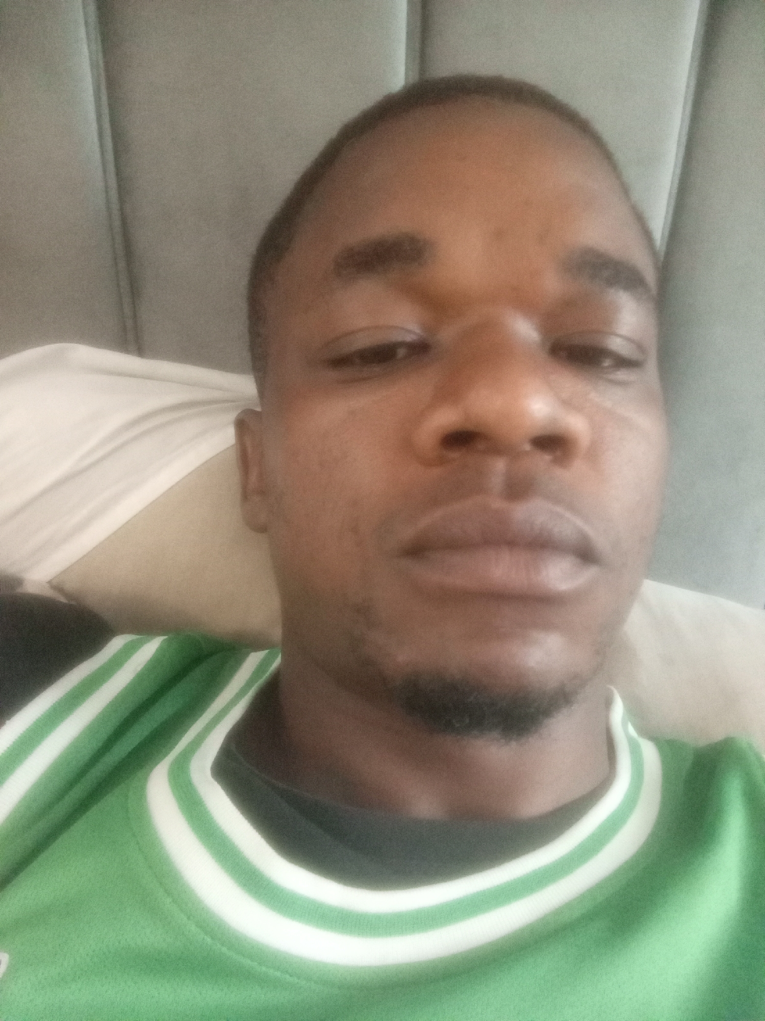 A close-up headshot of a dark-skinned man is shown. He has a short haircut and a neatly trimmed beard. The man is wearing a green sports jersey with white stripes around the neck. He appears to be lying down, possibly on a pillow, with a padded headboard visible in the background. There is no visual indication of a specific location in Kubwa, Nigeria.