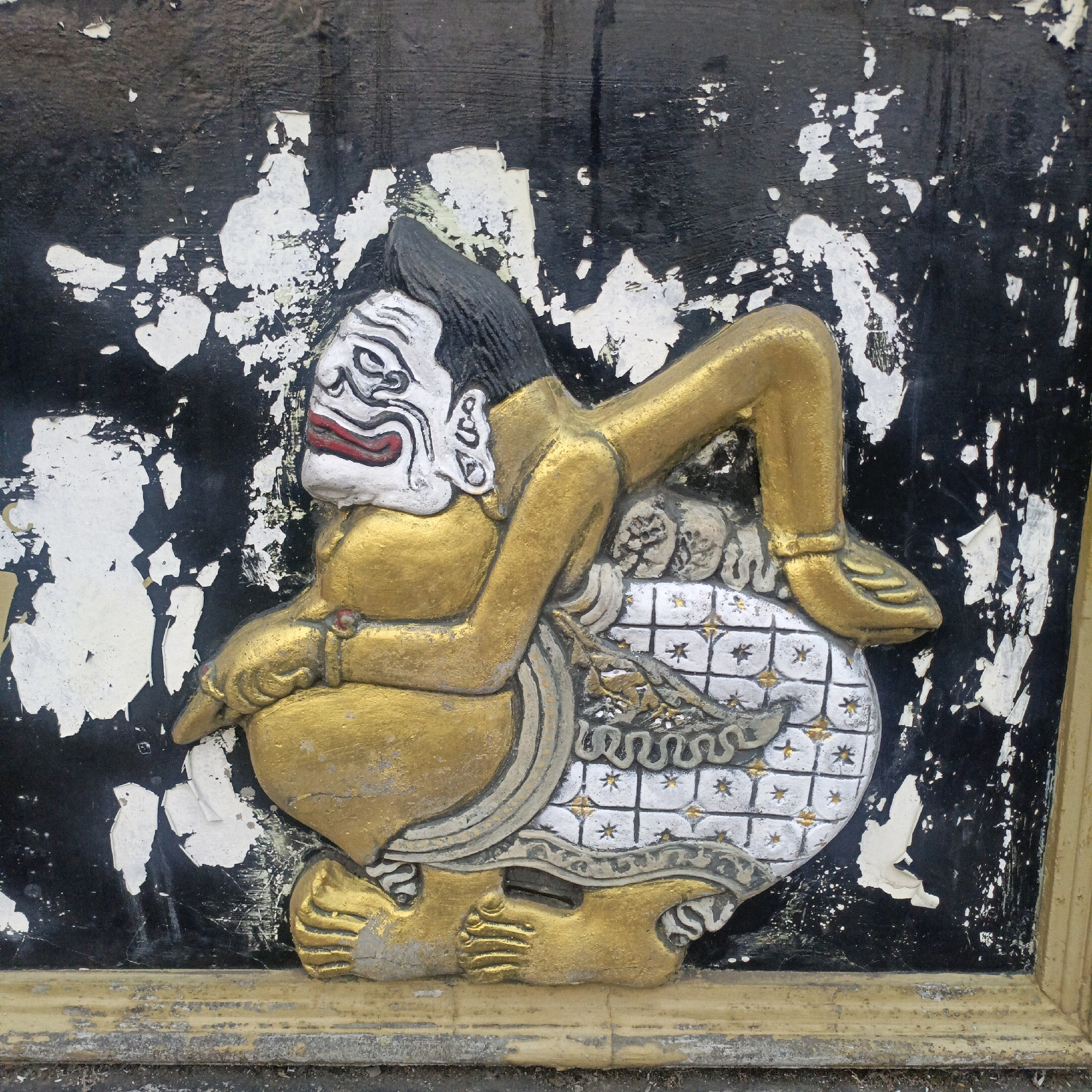 The image shows a close-up, eye-level view of a relief carving of a traditional Javanese puppet figure, possibly from a Wayang Kulit performance, or a similar cultural representation. The carving is mounted on a dark, textured background that appears to be wood or plaster with significant peeling and chipping of white paint, revealing the dark surface beneath. The figure itself is rendered in relief, with raised elements that create a sense of depth.
The puppet is predominantly painted in metallic gold, with accents of white, black, and red. The face is white with exaggerated features: black, arched eyebrows, wide, black-rimmed eyes with red accents, and a large, open mouth with red lips outlined in black. The figure has black hair that is slicked back. The body is a rich gold, with exaggerated proportions. It is depicted in a seated or reclining position, with one leg bent and extended, and the other foot resting on a patterned surface. The patterned surface appears to be a quilted or tiled cushion in white with a repeating pattern of small gold star-like motifs and some darker grey detailing.
The figure's arms are bent, with one hand resting on its chest and the other positioned near its lap. The carving shows intricate details in the clothing or ornamentation around the figure's waist, featuring flowing grey and white patterns with gold accents. The overall impression is of a stylized, theatrical character.
The context provided states the image is from Salatiga, Indonesia. This suggests the carving is likely a decorative element on a building, a sign, or part of a cultural display in that city. The condition of the background suggests it might be an outdoor or weathered installation. The style of the puppet strongly points to Indonesian cultural heritage.
