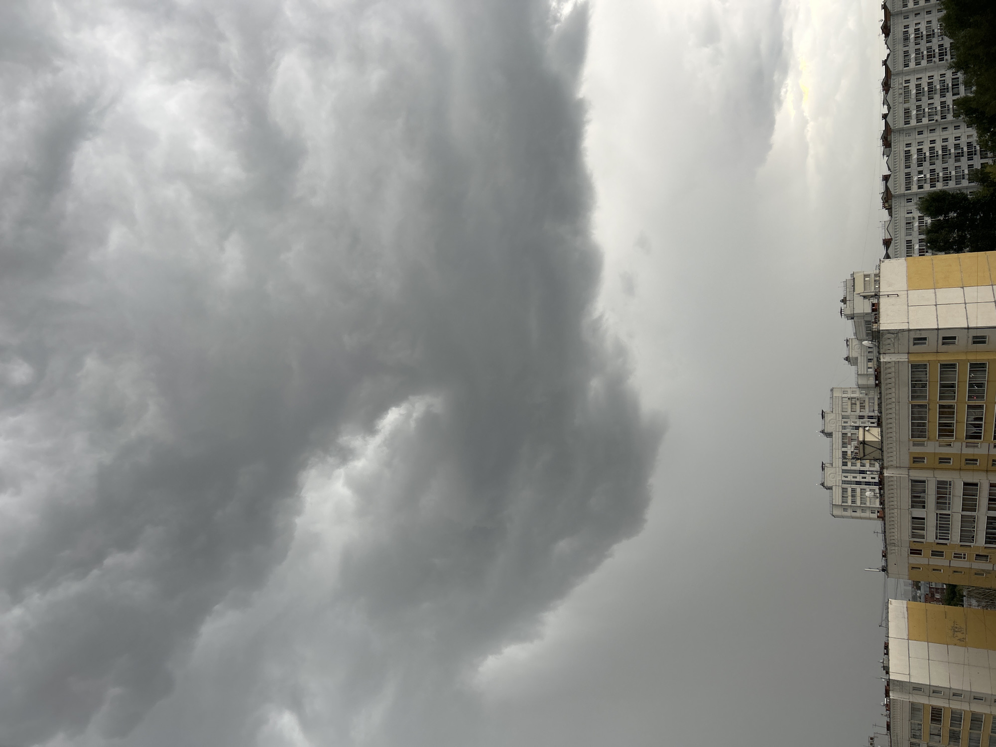 The image shows a view of the sky above a residential area in Tomsk, Russia. Dark, heavy storm clouds dominate the upper portion of the frame, obscuring much of the sky. Below the clouds, several multi-story apartment buildings are visible, characterized by their light-colored facades and numerous windows. Some of these buildings have yellow accents on their lower levels. Trees are interspersed between the buildings, and the overall atmosphere suggests impending rain or an ongoing weather event. There are no people or animals depicted in the image.