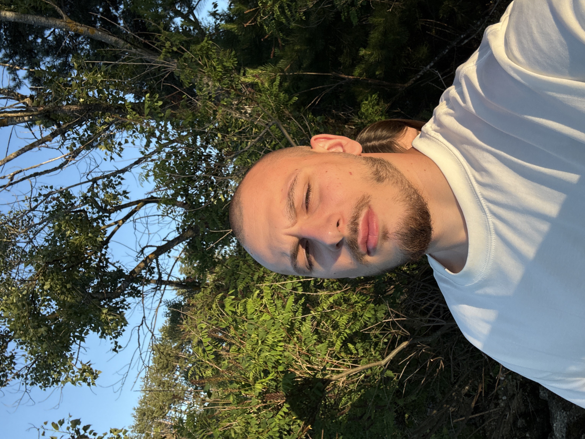 In Koprinka, Bulgaria, a young man with a shaved head and a beard takes a selfie outdoors during the day. He is wearing a white t-shirt and stands in front of lush green bushes and trees, with the blue sky visible above. Sunlight casts shadows on the foliage and highlights the man's face. A portion of another person's head and hair is visible behind the man on the right side of the frame. The scene suggests a casual outdoor setting, possibly a park or natural area.