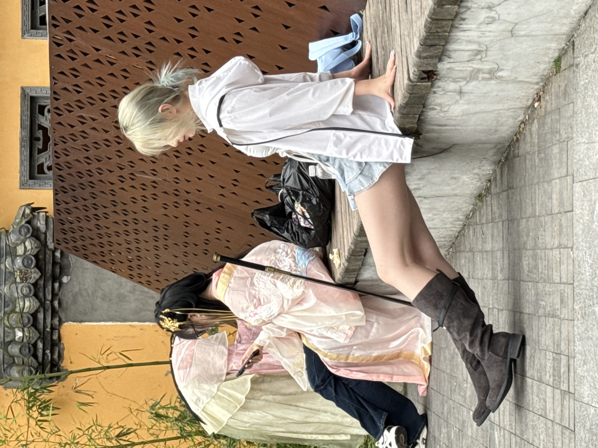 This outdoor photograph features two individuals resting on a wooden ledge, set against a backdrop blending traditional and modern architectural elements, likely in Yuzhong District, China. The scene appears to be taking place during clear daylight hours.
On the left, a person with blonde or light-colored hair, styled in a ponytail with faint blue streaks, is dressed in a white long-sleeved jacket with black trim and light blue denim shorts. Their hands, featuring long, white-tipped fingernails, are placed on the wooden ledge. A black bag sits on the paved ground nearby, and a light blue item rests on the ledge next to them.
To their right, another person wears traditional-style clothing, consisting of a light pink top with intricate blue and gold embroidery, including a dragon motif. Their dark hair is adorned with golden accessories. They wear dark blue jeans and white and black sneakers beneath the traditional garment. This individual holds a black smartphone in one hand and a slender, dark stick-like prop, possibly a sword hilt, in the other. They also wear dark brown, knee-high boots.
The background behind the left person features a reddish-brown wall with numerous triangular perforations, suggesting a modern design. Behind the right person, traditional Chinese architectural elements are visible, including a yellow-ochre wall, grey tiled roofing, and green bamboo stalks. The ground beneath them is a paved surface made of stone or concrete slabs. The overall mood is casual and relaxed. No visible text is present in the image.