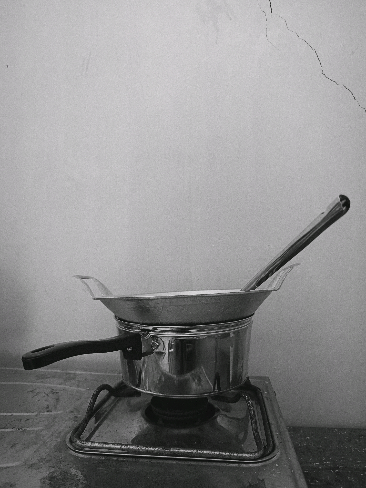 A black and white photograph depicts a stovetop with cooking utensils in what appears to be a kitchen or outdoor cooking area. A stainless steel pot sits on the stovetop, topped with a wok. A long-handled metal spatula or stirring utensil rests within the wok. The background is a textured wall with visible cracks. The stovetop is aged and shows signs of use. No people are present in the image. The scene is static and appears to be an indoor setting based on the wall texture and crack, but could potentially be an outdoor cooking setup given the simplicity of the stove. The image is devoid of any specific markers indicating its location in Panggungan, Indonesia.