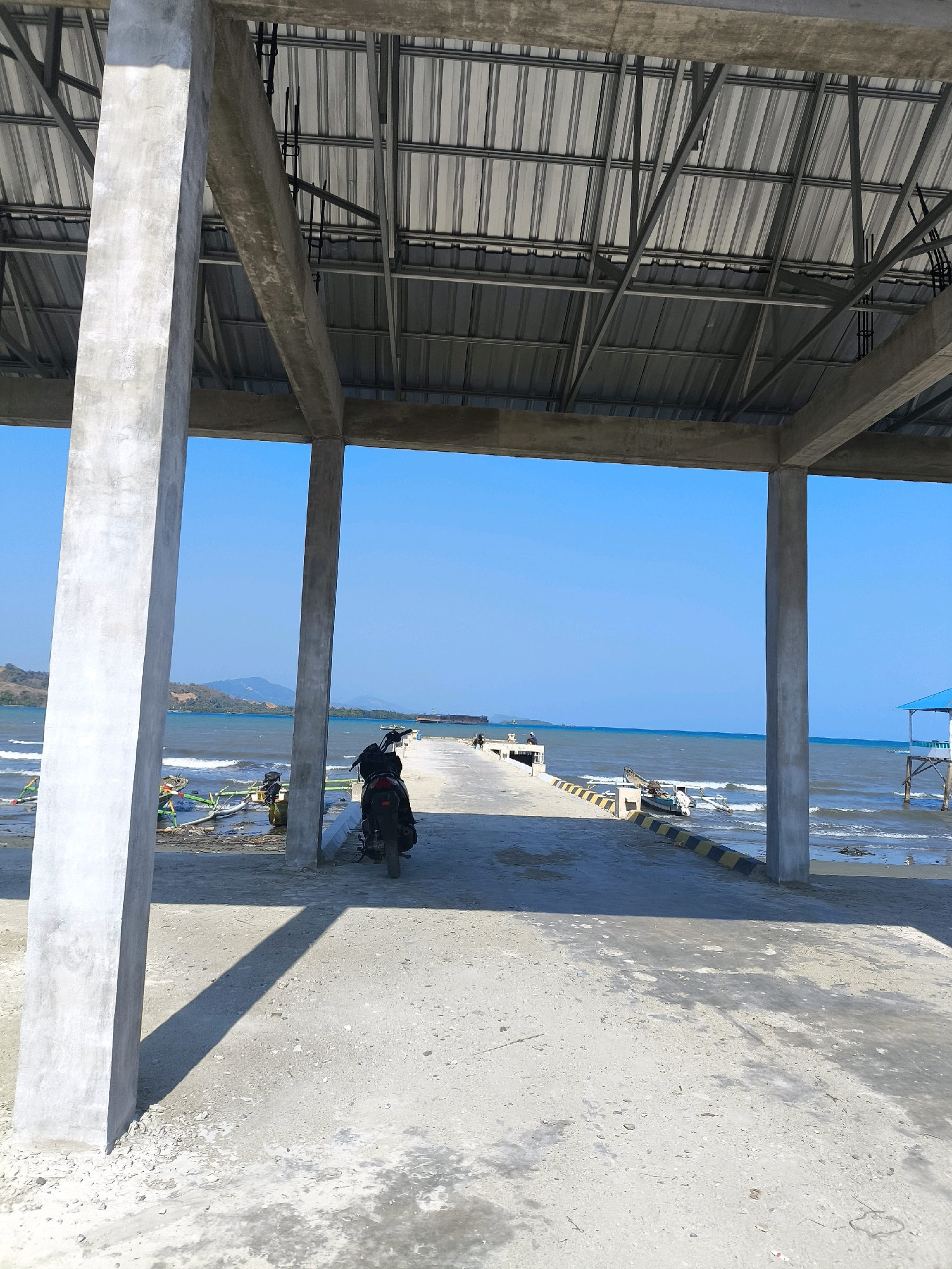 The image depicts a view from under a covered structure, likely a pier or a shelter, looking out towards the sea in Tilamuta, Indonesia. The structure features concrete pillars and a corrugated metal roof. In the foreground, a dark-colored motorcycle is parked. The ground is paved with concrete. Beyond the covered area, a concrete pier extends into the ocean. Small boats are visible docked along the pier and in the water. In the distance, the sea stretches to the horizon under a clear blue sky, with a faint outline of hills on the left. There are a few figures on the pier, suggesting human activity. The environment appears to be a coastal area, possibly a fishing port or a disembarkation point.