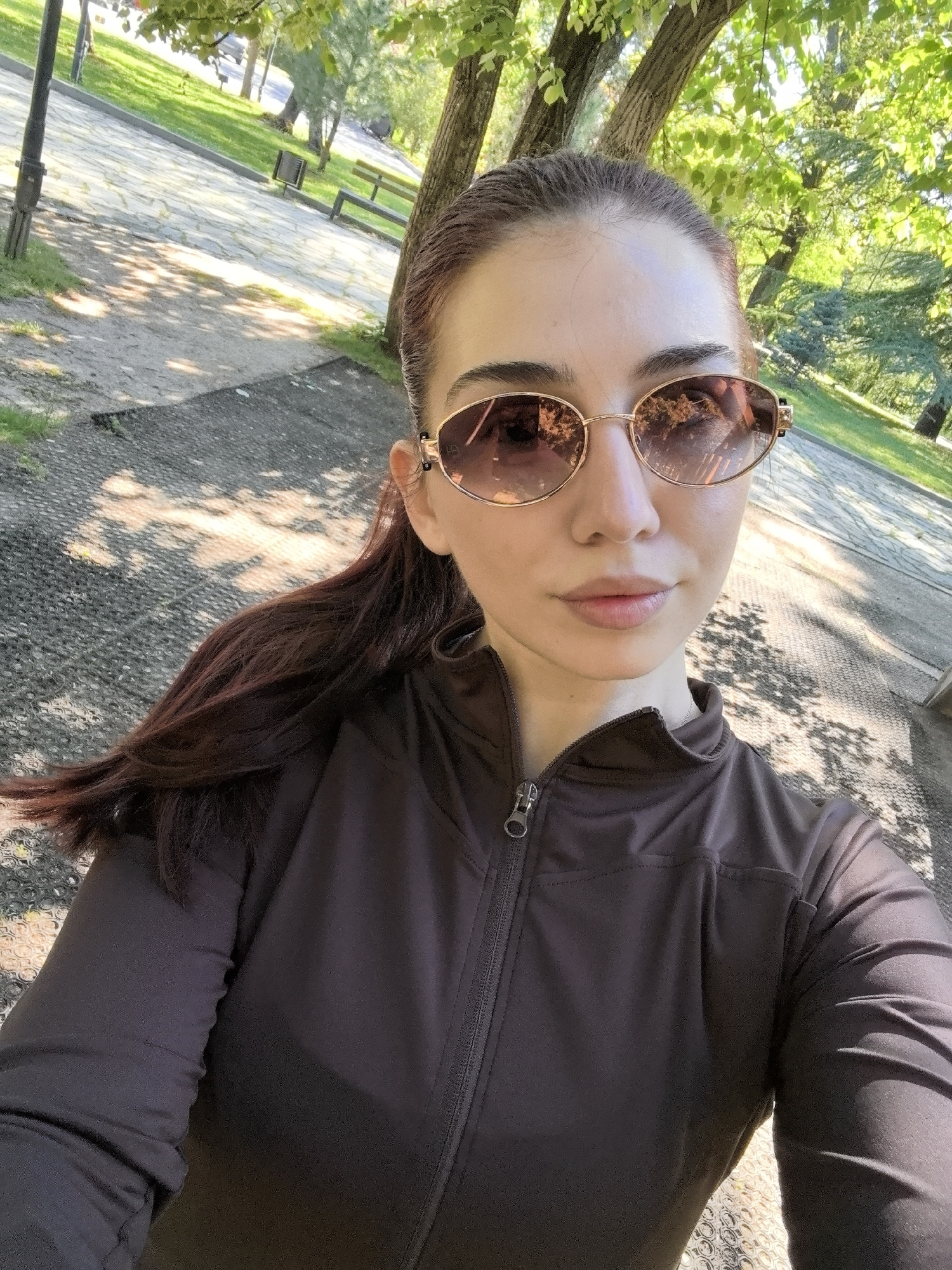 The image is a selfie of a woman taken outdoors in a park, likely in Stara Zagora, Bulgaria based on the vegetation, architecture, and context clues like public benches and landscaping typical of Bulgarian urban parks. The woman has reddish-brown hair pulled back in a ponytail and is wearing gold-rimmed sunglasses and a dark brown zippered jacket. The background shows a paved area with trees, suggesting a park walkway or plaza. There are benches and green spaces visible beyond the immediate foreground. The light indicates it is daytime. The woman is the primary subject, and her action is taking a selfie.