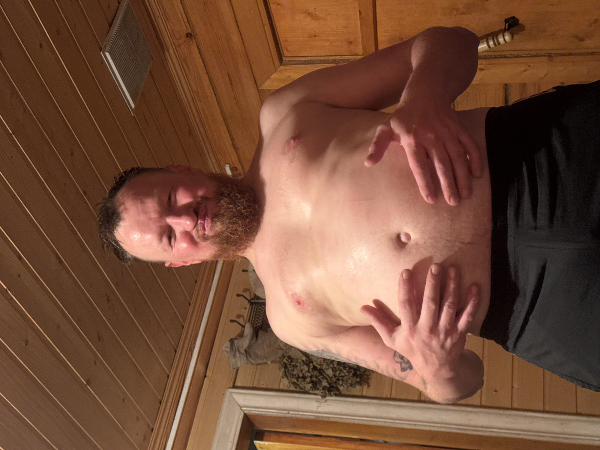 A shirtless man with a beard and graying hair stands in a room with wooden paneled walls, possibly a sauna. He is wearing dark shorts or pants. His skin appears sweaty. His hands are positioned on his torso. A ventilation grill is visible near the ceiling. A wooden rod is attached near the ceiling. A doorway and decorative elements are seen at the bottom. Based on the wooden paneling and sauna elements, the location might be a sauna or bathhouse. There is no contextual information linking the image to the city of Mayak, Russia.