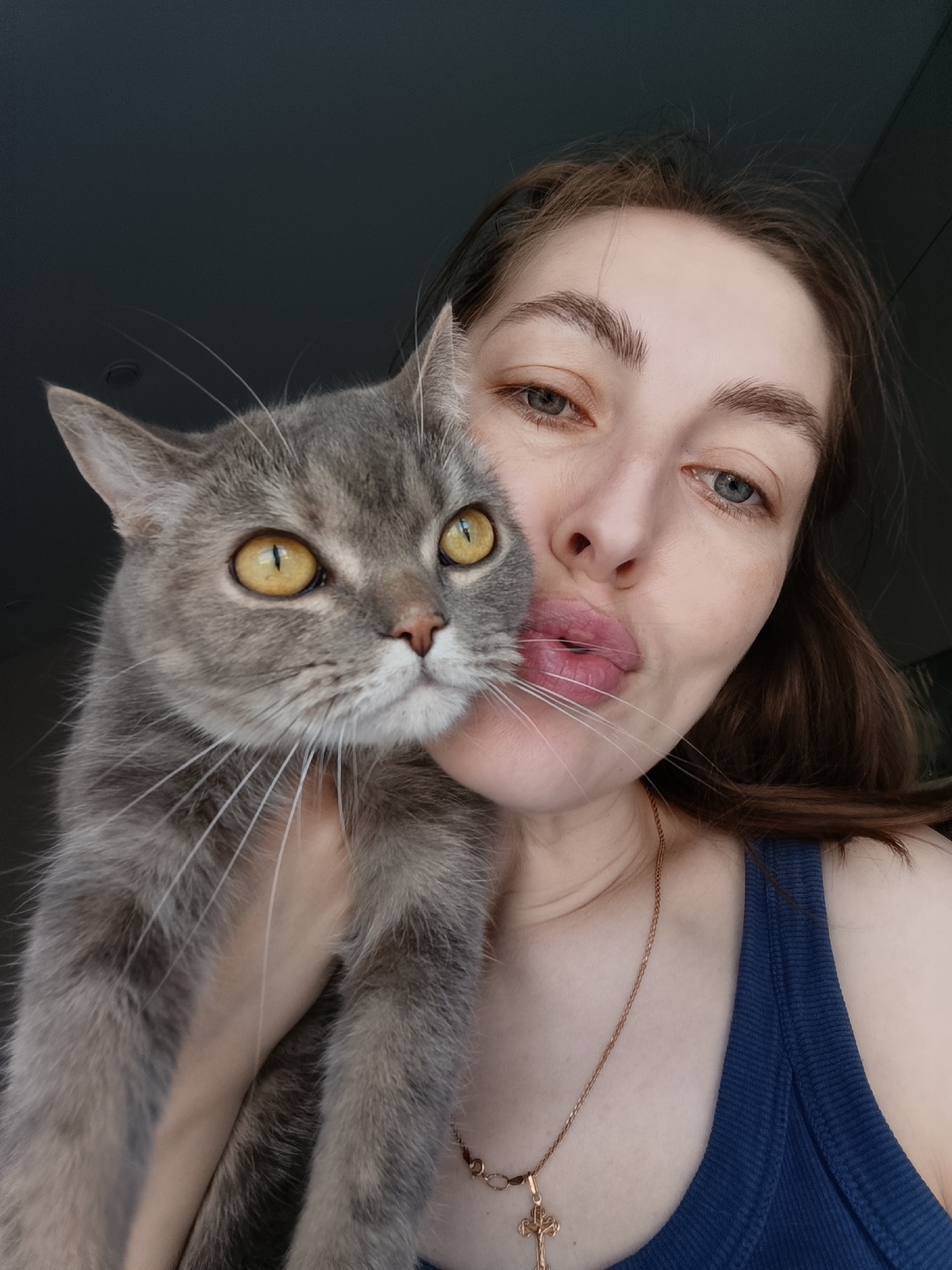A woman is holding a gray cat in front of her. The woman has light skin, brown hair, and blue eyes. She is wearing a blue tank top and a gold necklace with a cross pendant. She is making a kissing face at the camera. The cat has yellow eyes and a gray coat. It is looking to the left. The background is dark. The image was taken in the city of Chaikovsky, Russia.